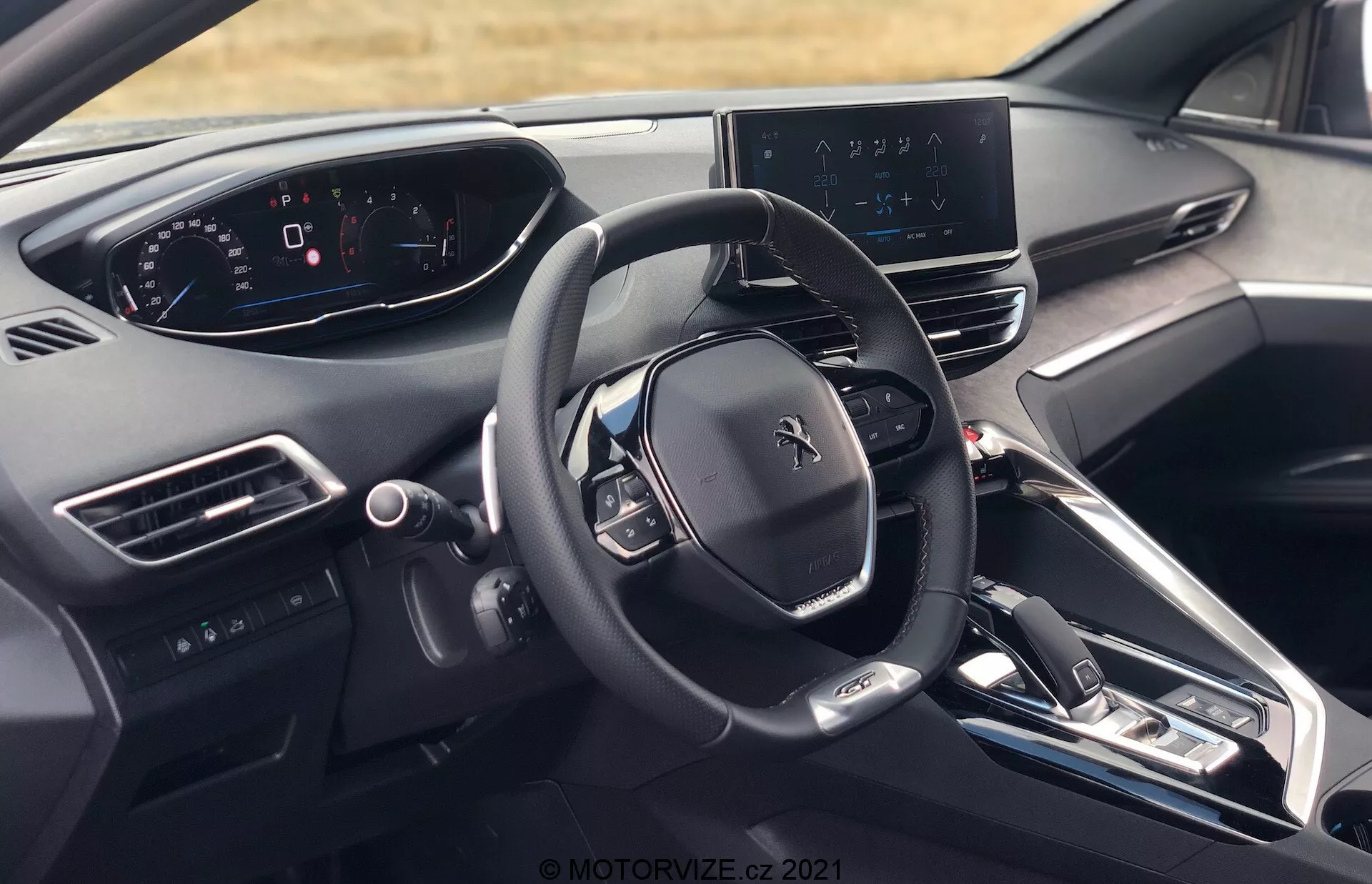 Vista interna della Peugeot 5008 2021 dalla prospettiva del conducente, che mostra il volante con il logo Peugeot, diversi pulsanti di controllo, quadro strumenti completamente digitale, display infotainment centrale con mappa di navigazione, cruscotto elegante con pochi pulsanti fisici, console centrale con pulsanti per il controllo del clima e le impostazioni del veicolo, leva del cambio elettronica, prese d'aria, pulsante di avviamento/arresto motore, materiali di alta gamma e vista parziale del poggiatesta del sedile del conducente.