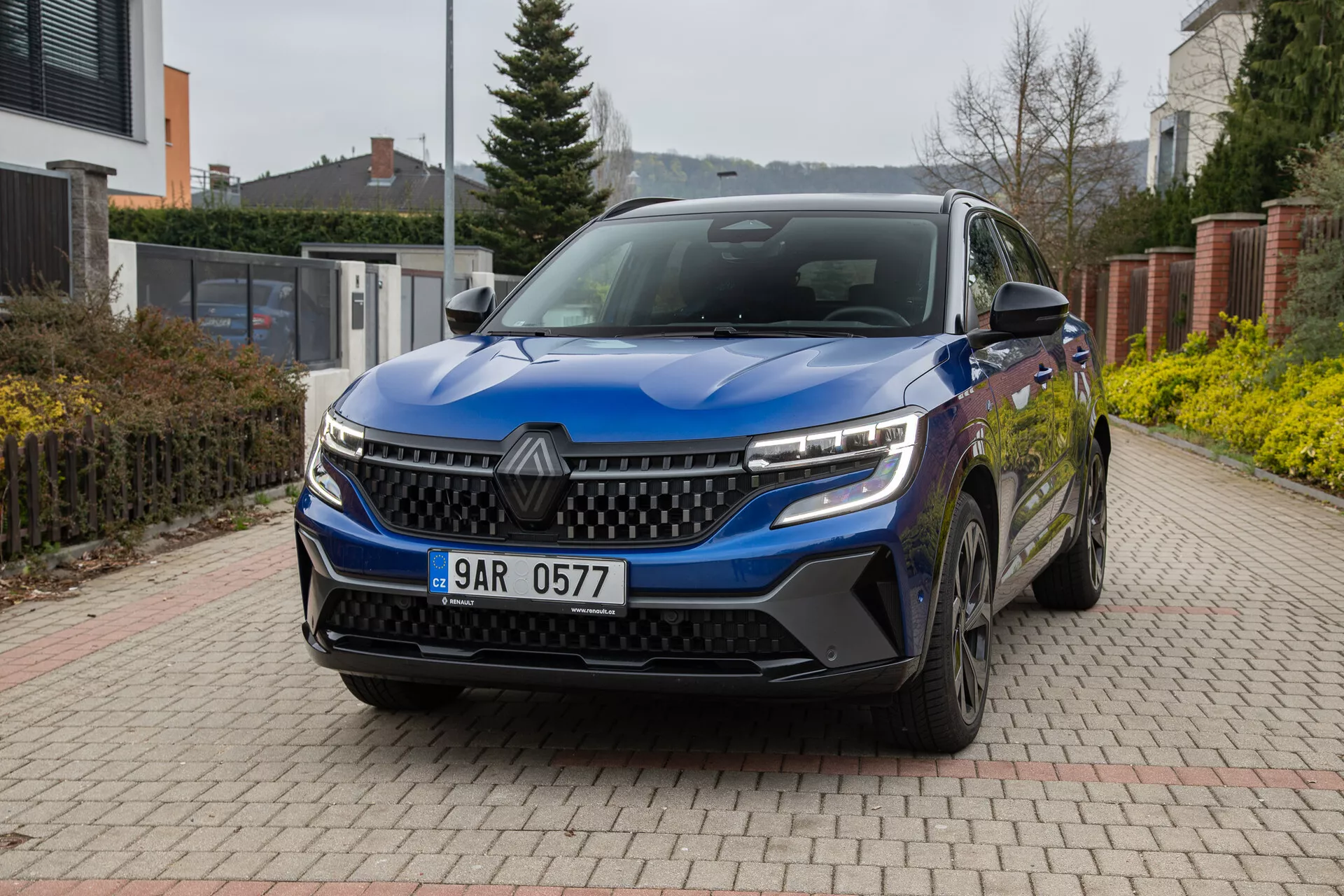 Renault Austral 2022 di colore blu, in mostra dal suo angolo anteriore a tre quarti su un vialetto pavimentato in mattoni con ambiente residenziale sullo sfondo, dotato di fari LED, griglia distintiva, sensori anteriori, cerchi in lega e specchietti laterali con indicatori di direzione integrati sotto una luce naturale nuvolosa.