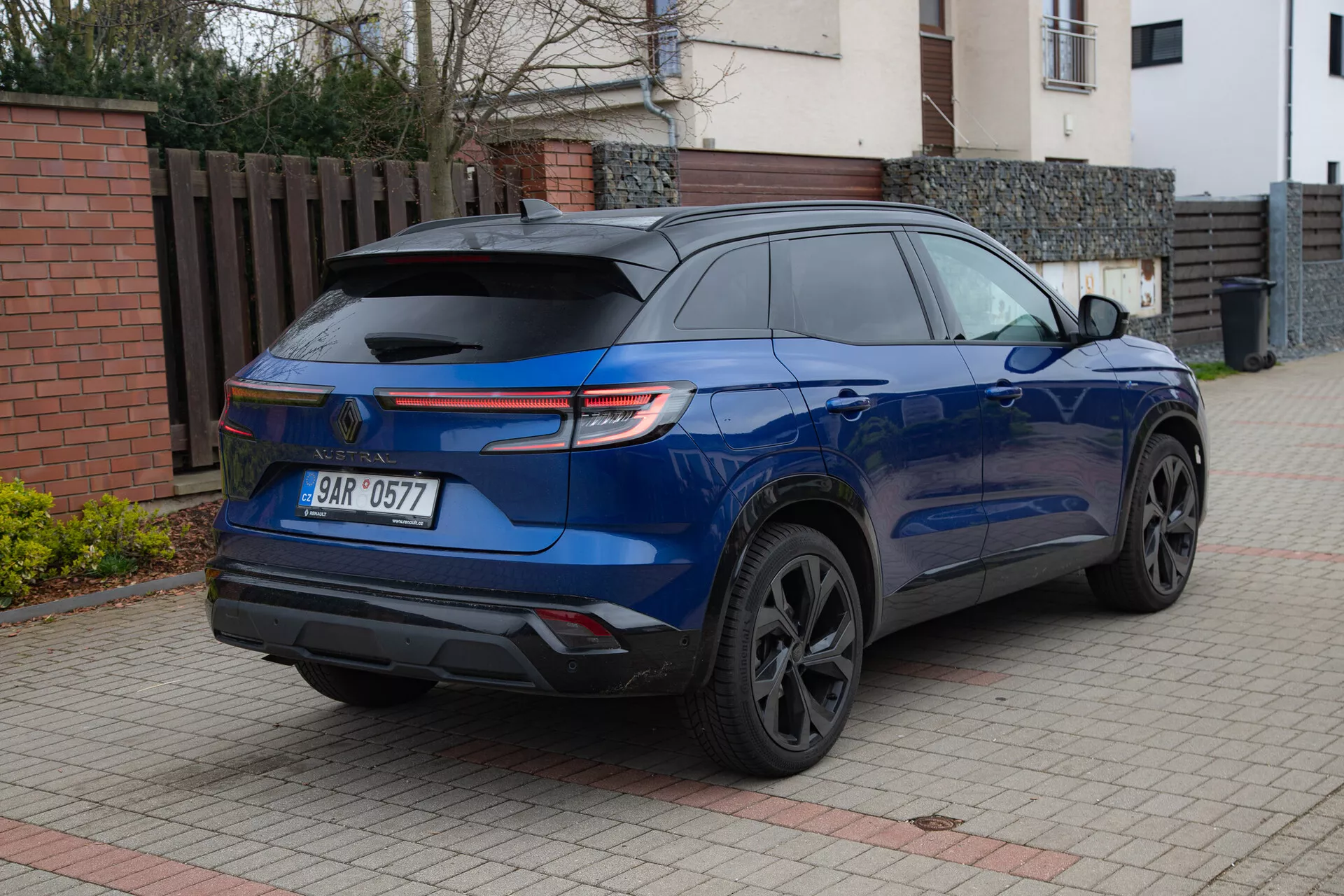 SUV moderno Renault Austral 2022 in blu metallico lucido parcheggiata su un vialetto pavimentato accanto a un edificio residenziale, mostrando il suo spoiler posteriore sportivo, fanali posteriori LED, cerchi in lega scuri, vetri posteriori oscurati e sensori di parcheggio da un angolo posteriore di tre quarti