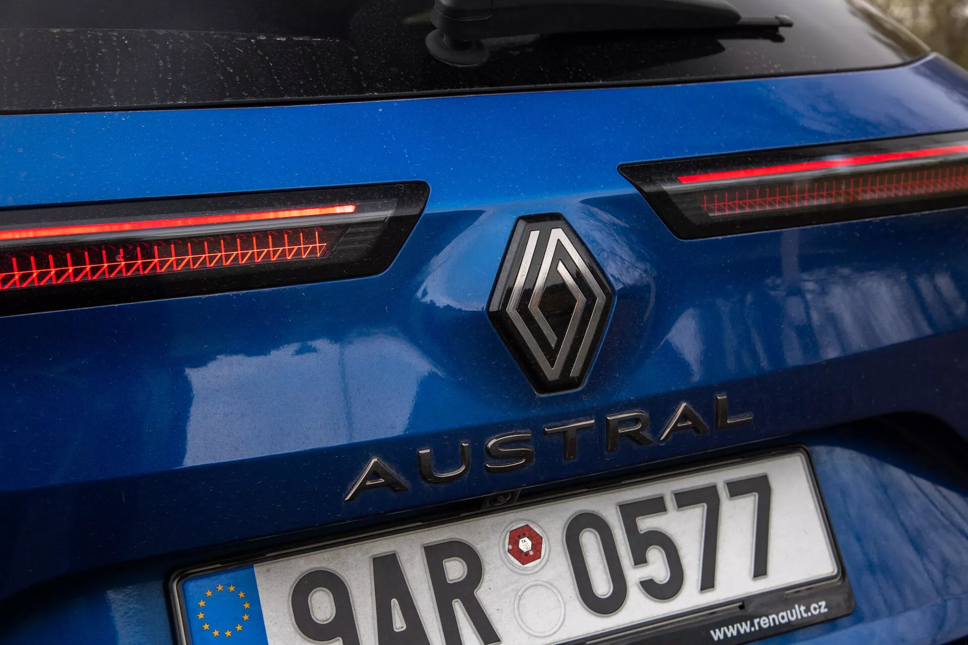 Immagine ravvicinata della sezione posteriore di una Renault Austral 2022 in blu lucido, con logo a diamante posizionato centralmente e nome del modello 'Austral' in lettere maiuscole, targa con bandiera dell'Unione Europea, distintivi fanali posteriori LED orizzontali e possibile ulteriore sistema di illuminazione posteriore, esterno dell'auto che indica un utilizzo recente.