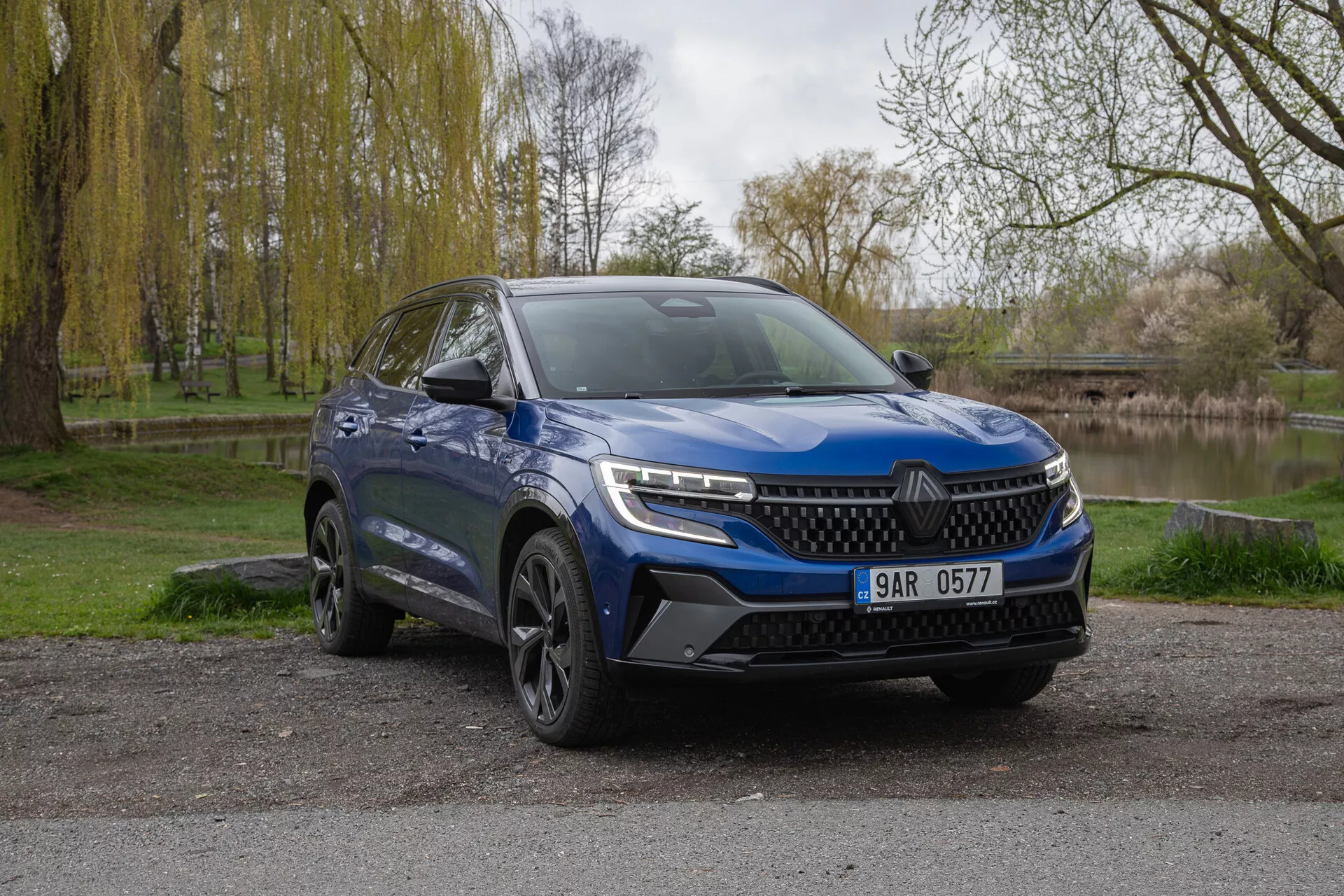 SUV blu Renault Austral 2022 parcheggiato all'aperto che mostra la facciata anteriore e il lato del guidatore con fari LED, paraurti sportivo e ruote in lega su sfondo naturale