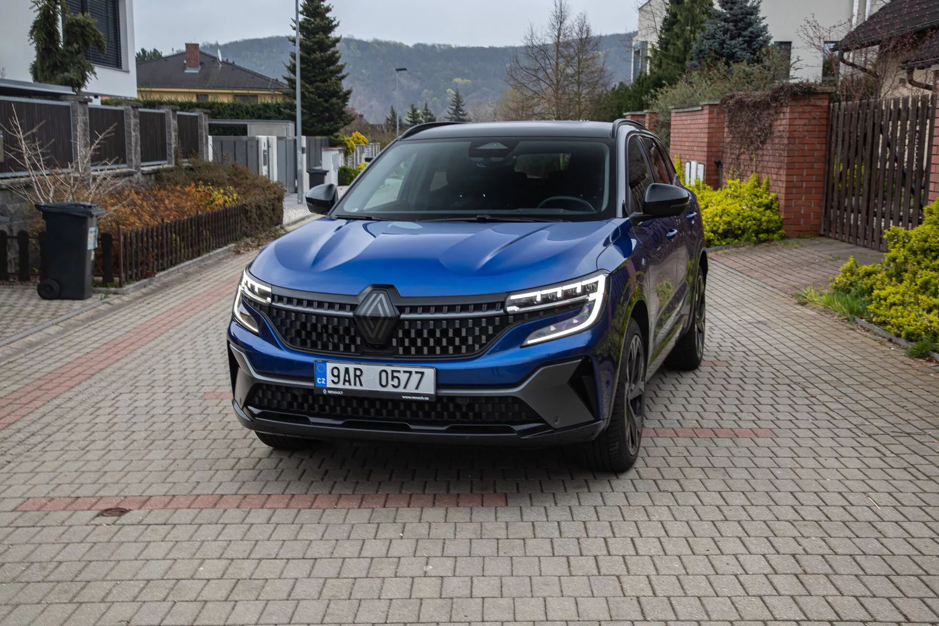 Vista frontale angolata di una Renault Austral 2022 SUV parcheggiata in un vialetto residenziale, mostrando la griglia, i fari LED, le luci diurne, il paraurti con prese d'aria, i sensori di parcheggio, l’emblema, le ruote in lega, gli specchietti laterali con indicatori di direzione, e la targa.