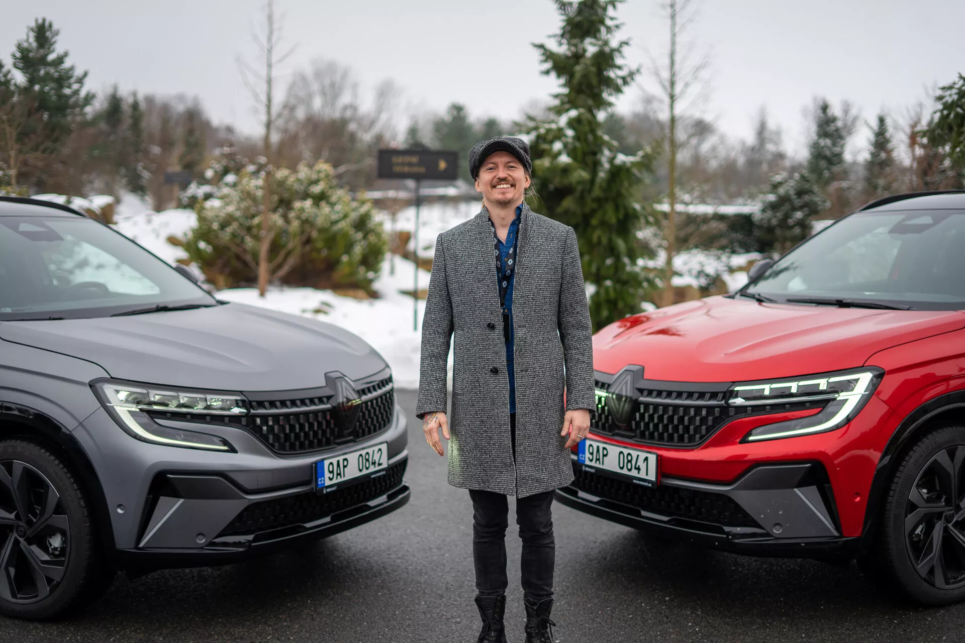 Persona sorridente affiancata da due SUV Renault Austral 2022 in un contesto invernale, che evidenzia il design moderno dei veicoli e le caratteristiche tecnologiche avanzate con un focus sulla griglia anteriore, fari LED e profilo laterale elegante.