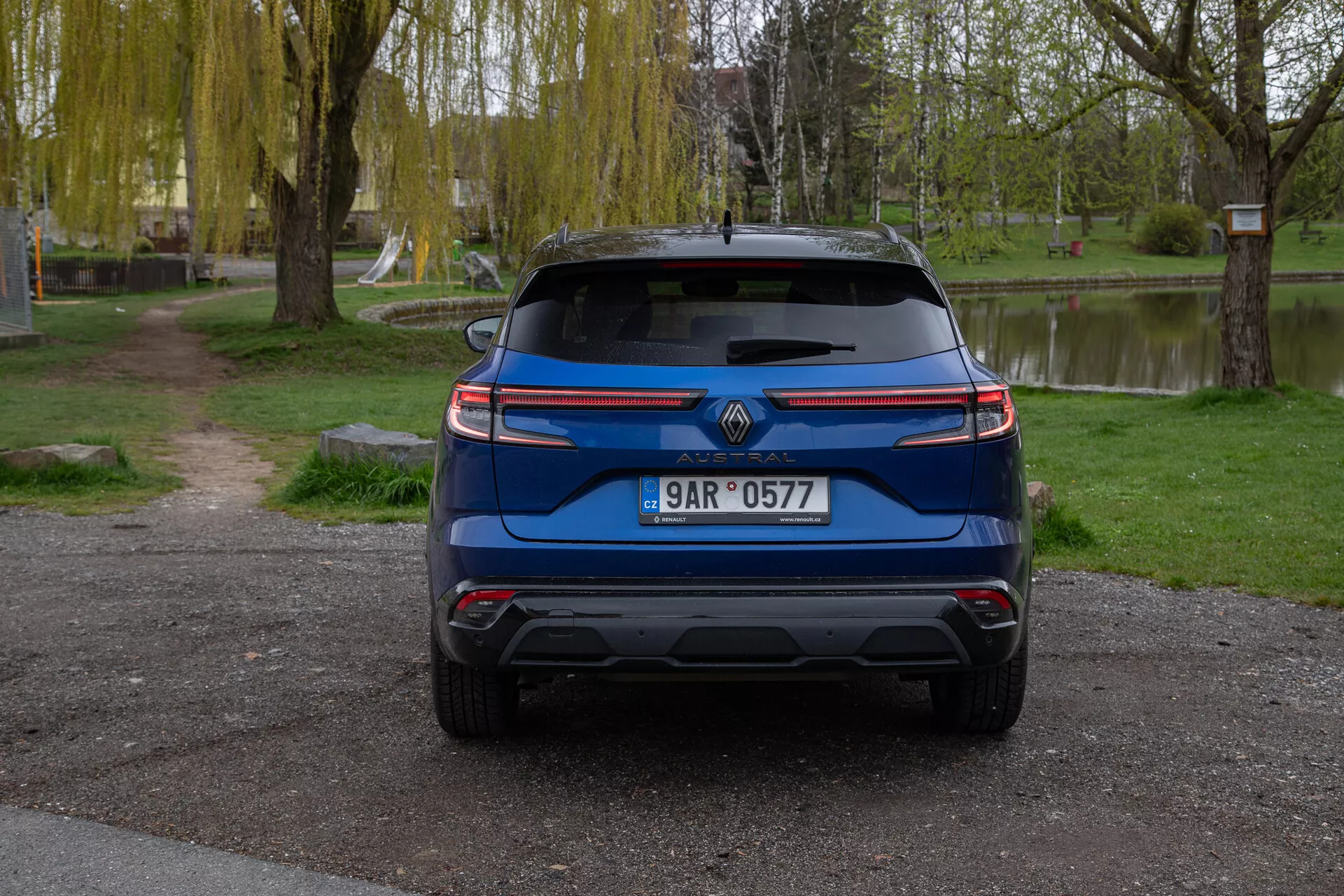 Vista posteriore di una Renault Austral 2022 blu, parcheggiata all'aperto mostrando caratteristiche che includono il logo del marchio, targa, luci posteriori moderne, parabrezza posteriore con tergicristallo, paraurti posteriore progettato con potenziali sensori di assistenza al parcheggio, punte di scarico visibili, riflettori o sensori, e uno spoiler posteriore per migliorare l'aerodinamica, il tutto ambientato contro un tranquillo sfondo simile a un parco con alberi, erba e un corpo d'acqua.