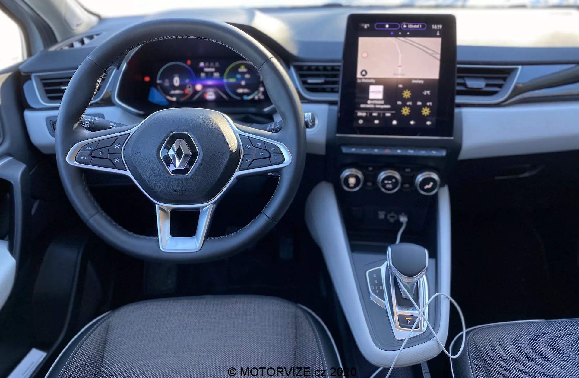 Inquadratura degli interni della Renault Captur 2020 dalla prospettiva del conducente, evidenziando il volante di design moderno con comandi multimediali integrati, il quadro strumenti digitale, il sistema di infotainment con schermo touch verticale e controlli HVAC, la leva del cambio automatico e la connettività USB. L'illuminazione e la qualità suggeriscono un interno ben progettato e tecnologicamente avanzato.