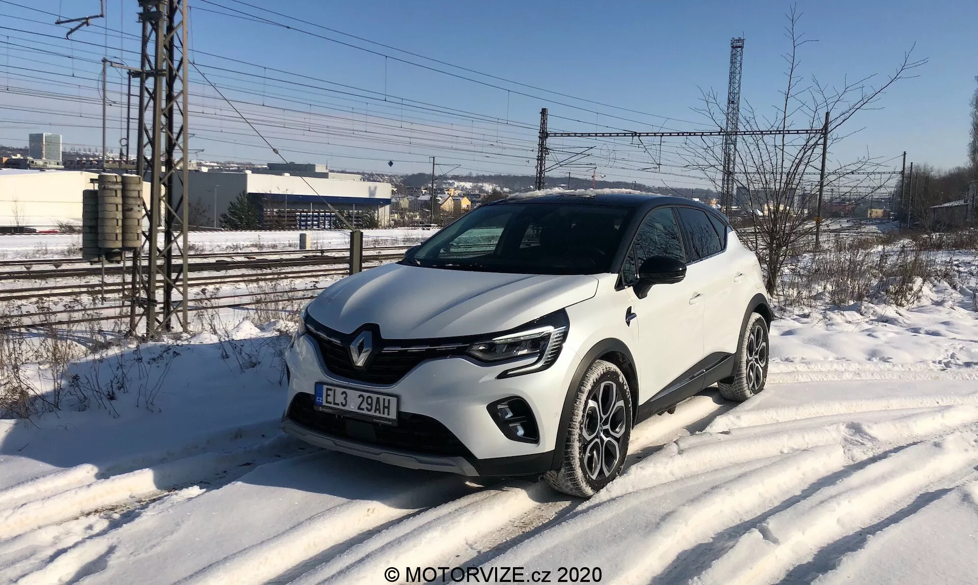 L'immagine mostra una Renault Captur 2020 bianca, un SUV compatto parcheggiato su un bordo strada coperto di neve, catturato da un'angolazione frontale di tre quarti. Il veicolo presenta luci diurne a LED distintive a forma di C, fari a proiettore, una griglia frontale con accenti neri con un prominente stemma Renault e cerchi in lega a più razze. Elementi di design degni di nota includono rivestimenti in plastica lungo i bordi inferiori e gli archi delle ruote. L'ambiente suggerisce una giornata fredda invernale con uno sfondo industriale o semiurbano.