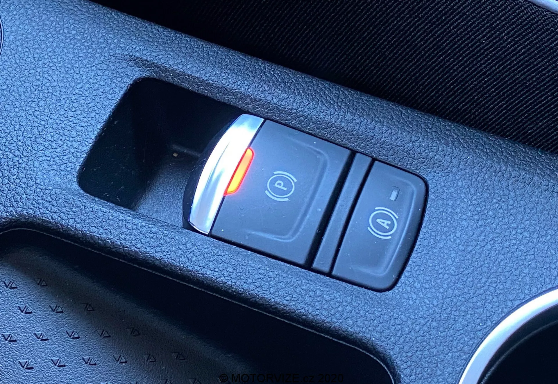 Primo piano diagonale dell'interruttore del freno di stazionamento elettronico nell'interno della Renault Captur 2020. L'interruttore, posizionato nella console centrale, presenta due pulsanti: uno illuminato di rosso con un lucchetto chiuso e il simbolo 'P' che indica lo stato attivato, e un altro pulsante con il simbolo del lucchetto aperto. L'interruttore è inserito in una console di plastica nera testurizzata, evidenziando la tecnologia moderna del veicolo.