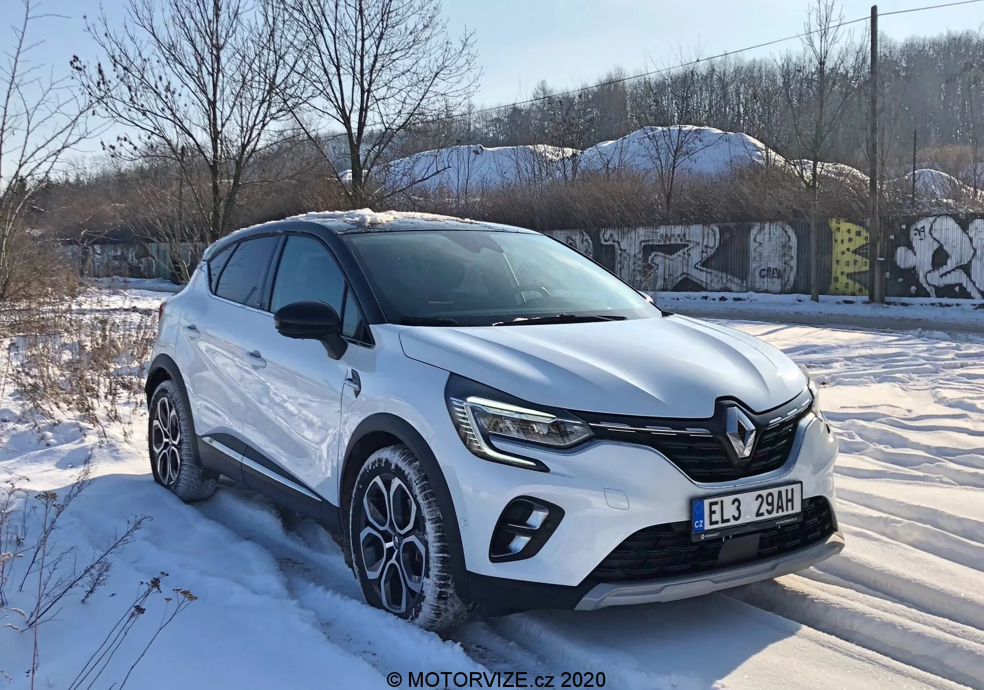 Renault Captur 2020 vista tre quarti frontale che mostra il design moderno del crossover con una distintiva griglia anteriore, fari a LED affilati, protezioni del corpo vettura, grandi cerchi in lega a più razze e colore della carrozzeria bianco sotto condizioni invernali nevose in un ambiente urbano.