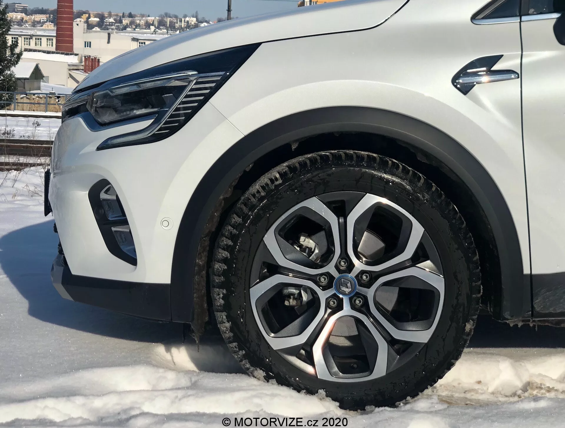 Primo piano di una Renault Captur 2020 da un angolo basso che mostra la sezione anteriore sinistra, con particolare attenzione alla ruota in lega, al parafango, al paraurti e all'area del faro. L'auto si trova su una superficie innevata in un contesto urbano con edifici e cielo sereno.