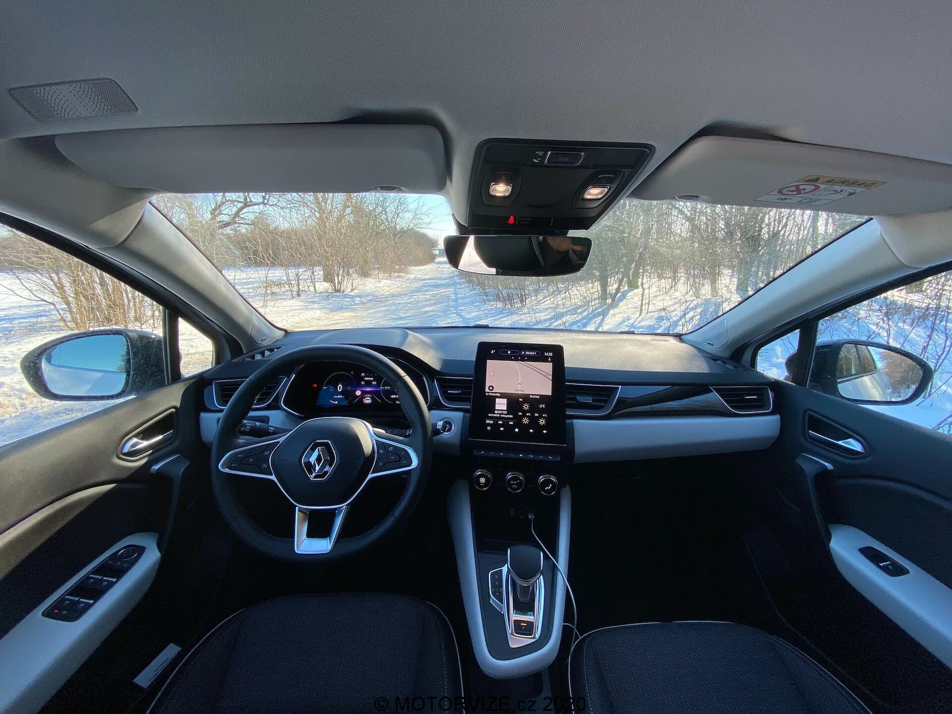 Immagine degli interni del Renault Captur 2020 dai sedili posteriori che mostra la cabina anteriore con volante a tre razze con pulsanti integrati, cluster digitale degli strumenti, sistema di infotainment touchscreen orientato verticalmente, console centrale moderna con pochi controlli fisici, selettore del cambio dal design elegante, bocchette di ventilazione HVAC, materiali premium inclusi pelle e accenti metallici, specchietto retrovisore probabilmente con sensori per ADAS e montante A visibile con comandi finestra.