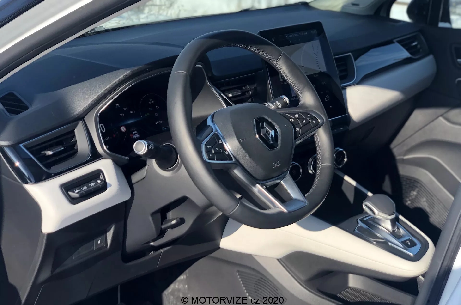 Vista interna della Renault Captur 2020 dalla prospettiva del conducente, con focus sul volante con il logo Renault, comandi montati, quadro strumenti digitale dietro e un sistema di infotainment touchscreen centralmente posizionato. L'immagine mette in risalto una combinazione di materiali sintetici e plastici con accenti metallici, trasmissione automatica, sistema di controllo del clima e regolazioni dei finestrini e degli specchietti elettrici.
