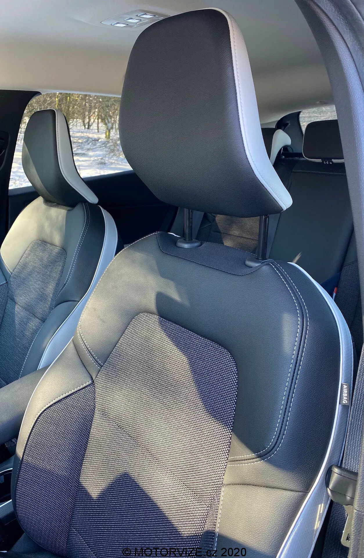 Vista interna della Renault Captur 2020 che mostra il sedile del passeggero anteriore e del conducente da un angolo posteriore, leggermente decentrato. La parte superiore dei sedili include poggiatesta integrati e telai visibili delle cinture di sicurezza. I sedili presentano un design moderno bicolore con colori grigio scuro e chiaro e bordi in pelle sintetica con dettagli cuciti, evidenziando artigianalità e stile.