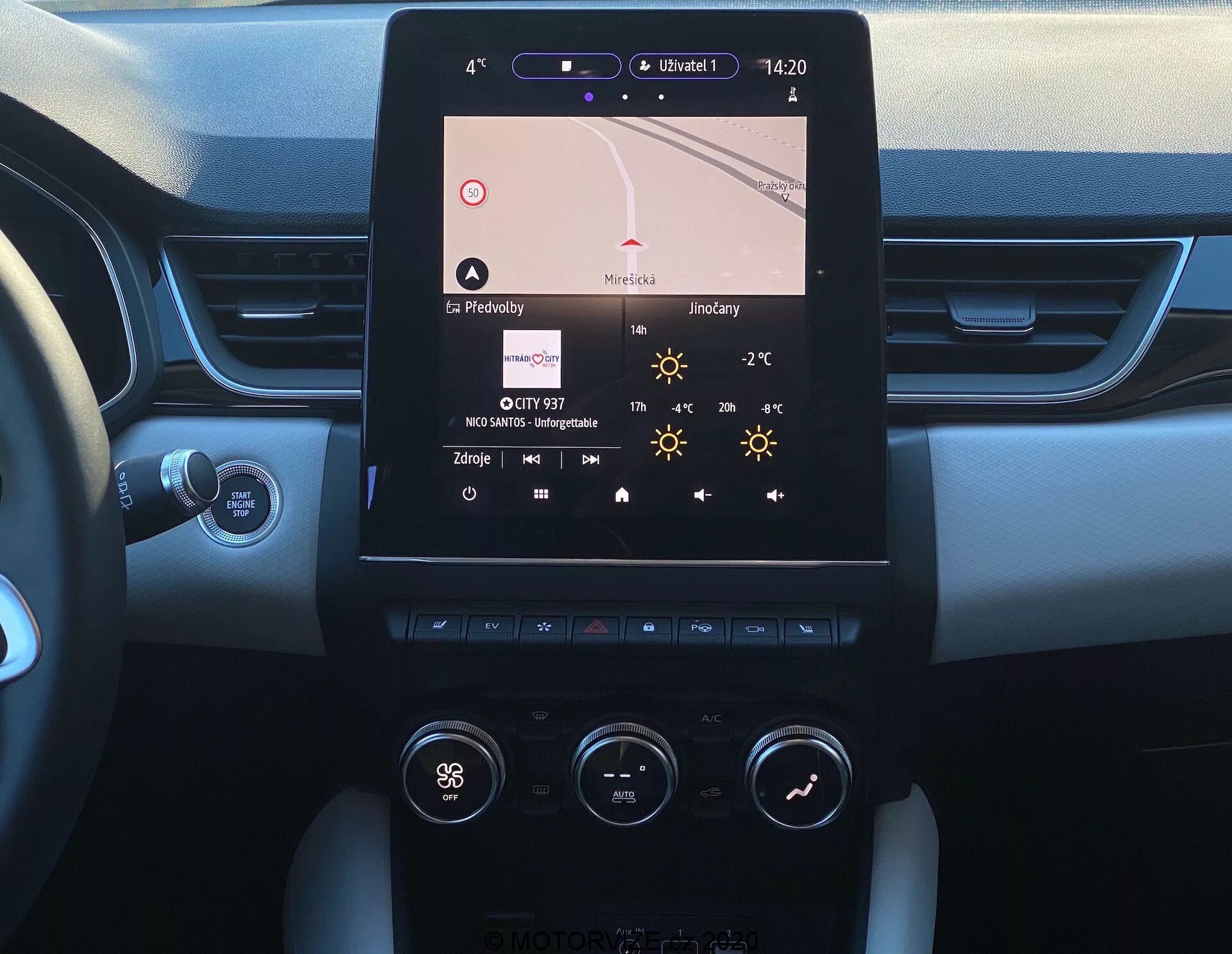 L'immagine mostra la consolle centrale di una Renault Captur 2020, vista dal sedile del conducente. La caratteristica principale è un display infotainment touchscreen orientato verticalmente che mostra la navigazione, l'ora (14:20), la temperatura (27°C) e i dettagli della radio. Sotto, è visibile il sistema di climatizzazione bizona con manopole e pulsanti che indicano impostazioni come 'Hi' per la temperatura del lato conducente. La consolle combina materiali metallici e plastici attorno allo schermo e ai controlli. I bordi del cruscotto rivelano un design bicolore e tattile morbido, e un pulsante Start/Stop è parzialmente visibile. Si vede la colonna dello sterzo con un'asta di controllo, probabilmente per i segnali di svolta o altre funzioni.