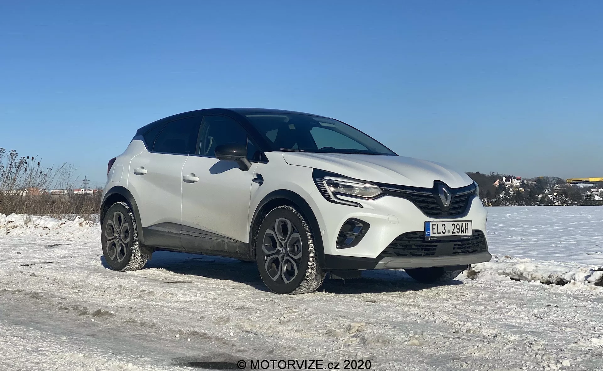 Vista a tre quarti di un SUV crossover compatto Renault Captur 2020 bianco in un ambiente invernale, con terreno coperto di neve e cielo azzurro limpido. L'immagine mette in evidenza il lato anteriore sinistro e parte del profilo laterale del veicolo, ripresa da un'angolazione leggermente inferiore che enfatizza le ruote e la metà inferiore. Il design moderno dell'auto mostra linee distinte sul cofano, fari LED eleganti con luci diurne e una griglia anteriore prominente. Le ruote in lega uniche e la plastica di protezione attorno ai parafanghi conferiscono un aspetto robusto. Le ombre sotto l'auto suggeriscono che la foto sia stata scattata a mezzogiorno o nel primo pomeriggio alla luce naturale del giorno.