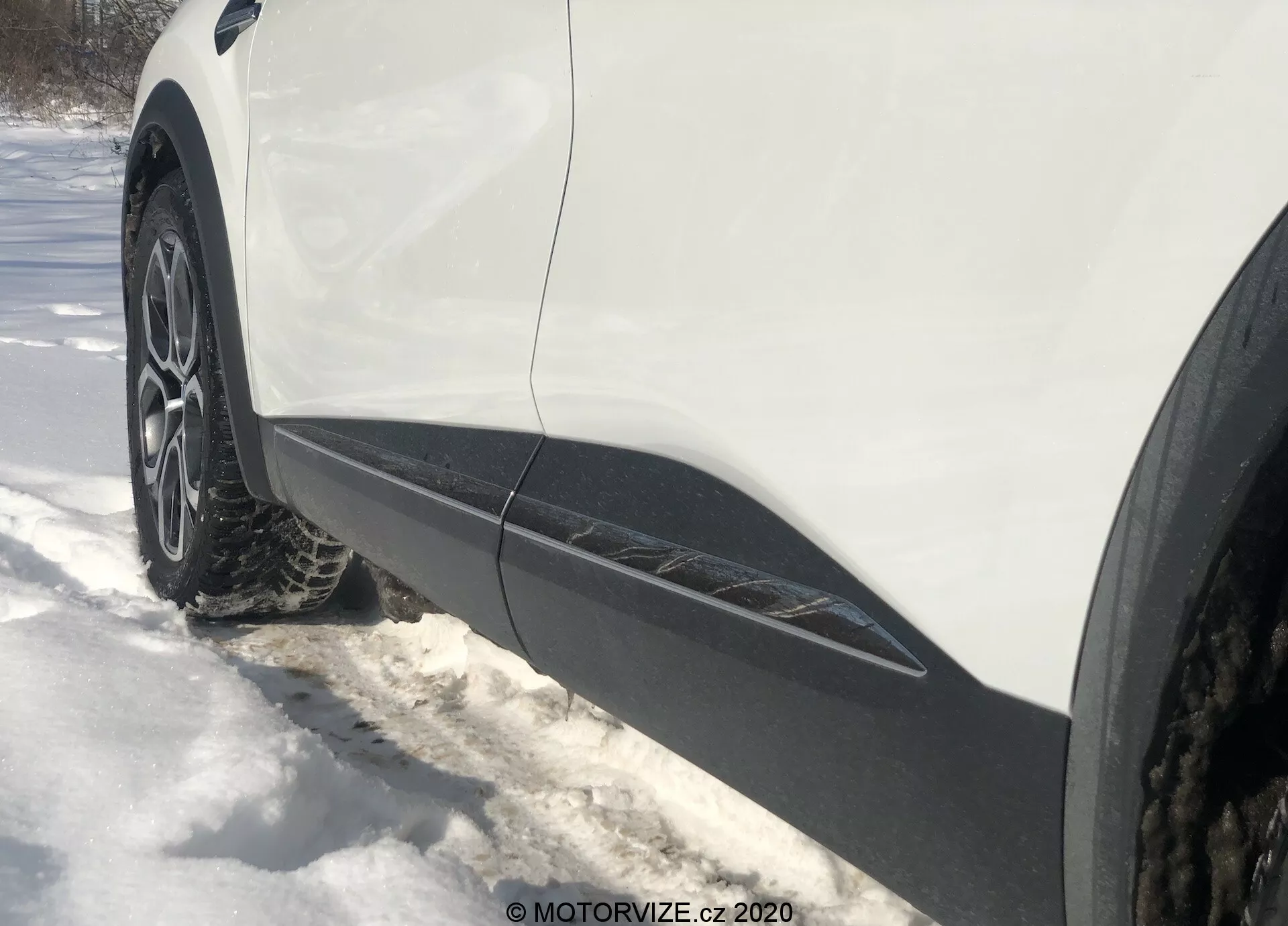 Vista laterale ravvicinata e angolata dal livello del suolo della ruota anteriore e del parafango lato guida di una Renault Captur 2020. Il cerchio in lega multi-razze con pneumatico invernale o per tutte le stagioni è circondato dalla neve. La carrozzeria bianca con finiture nere corre lungo il bordo inferiore, indicando un crossover o SUV. L'illuminazione suggerisce una giornata soleggiata con ombre visibili.