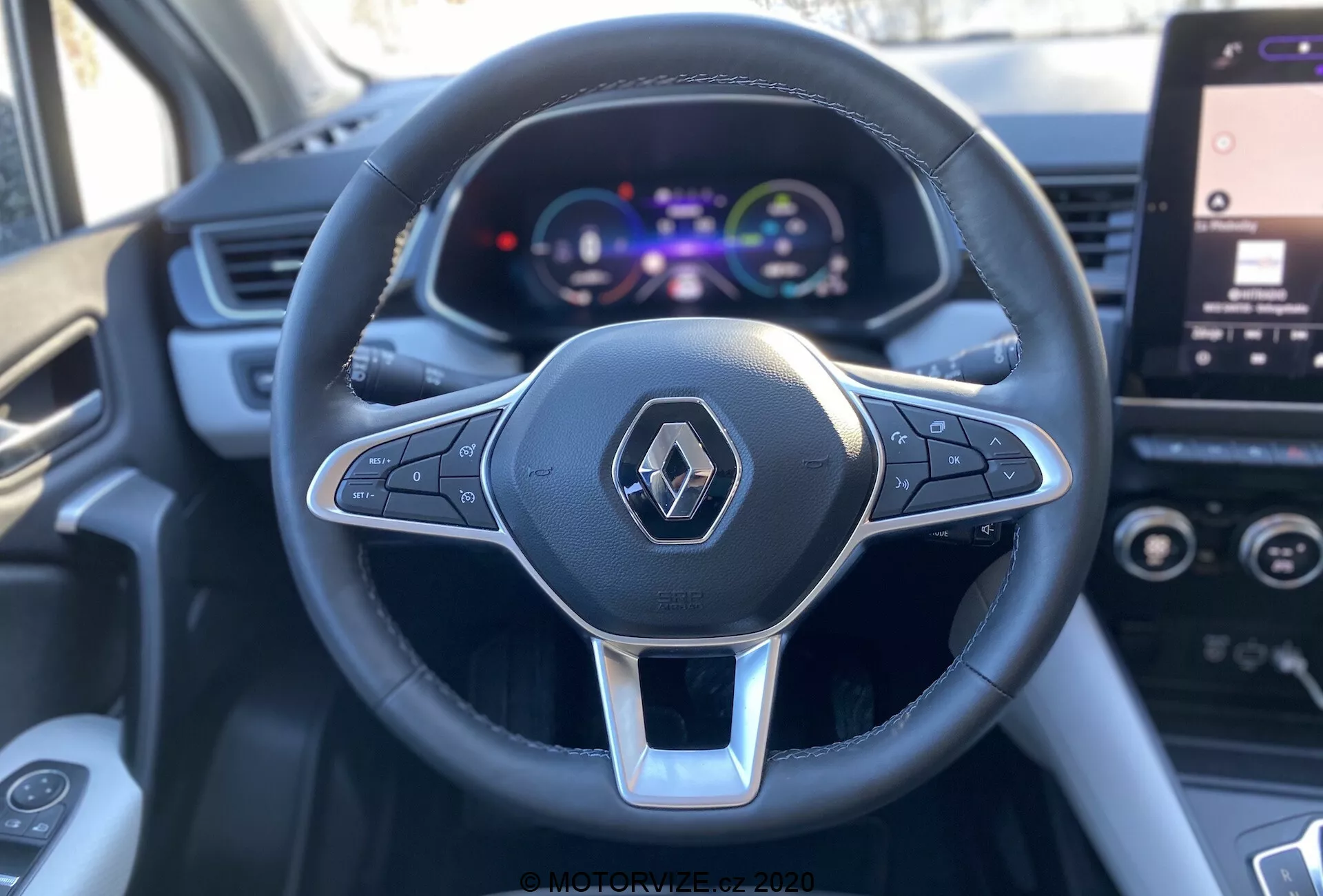 Vista ravvicinata in prima persona dal sedile del conducente di una Renault Captur 2020, focalizzata sul volante multifunzionale a tre razze rivestito in pelle con accento argentato e logo Renault, insieme a un cruscotto digitale a colori che mostra tachimetro e dettagli del veicolo, con un touchscreen di infotainment parzialmente visibile sullo sfondo.