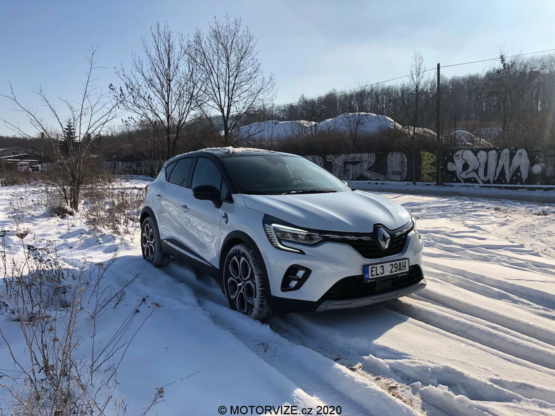 Un crossover SUV Renault Captur 2020 bianco è parcheggiato su un terreno innevato con vegetazione e strutture coperte di graffiti sullo sfondo, sotto un cielo per lo più sereno con alcune nuvole. L'immagine è scattata da un angolo frontale di tre quarti focalizzato sul lato del guidatore, mostrando la griglia anteriore con un emblema cromato e fari a LED contemporanei. Il SUV presenta luci diurne a LED anteriori, alloggiamenti per fari antinebbia, linee caratteriali sul cofano, rivestimenti in plastica intorno ai passaruota, specchietti laterali con indicatori di direzione integrati, cerchi in lega multi-razze con pneumatici invernali e una linea della spalla forte. La posizione elevata implica una maggiore altezza da terra, adatta a vari terreni.