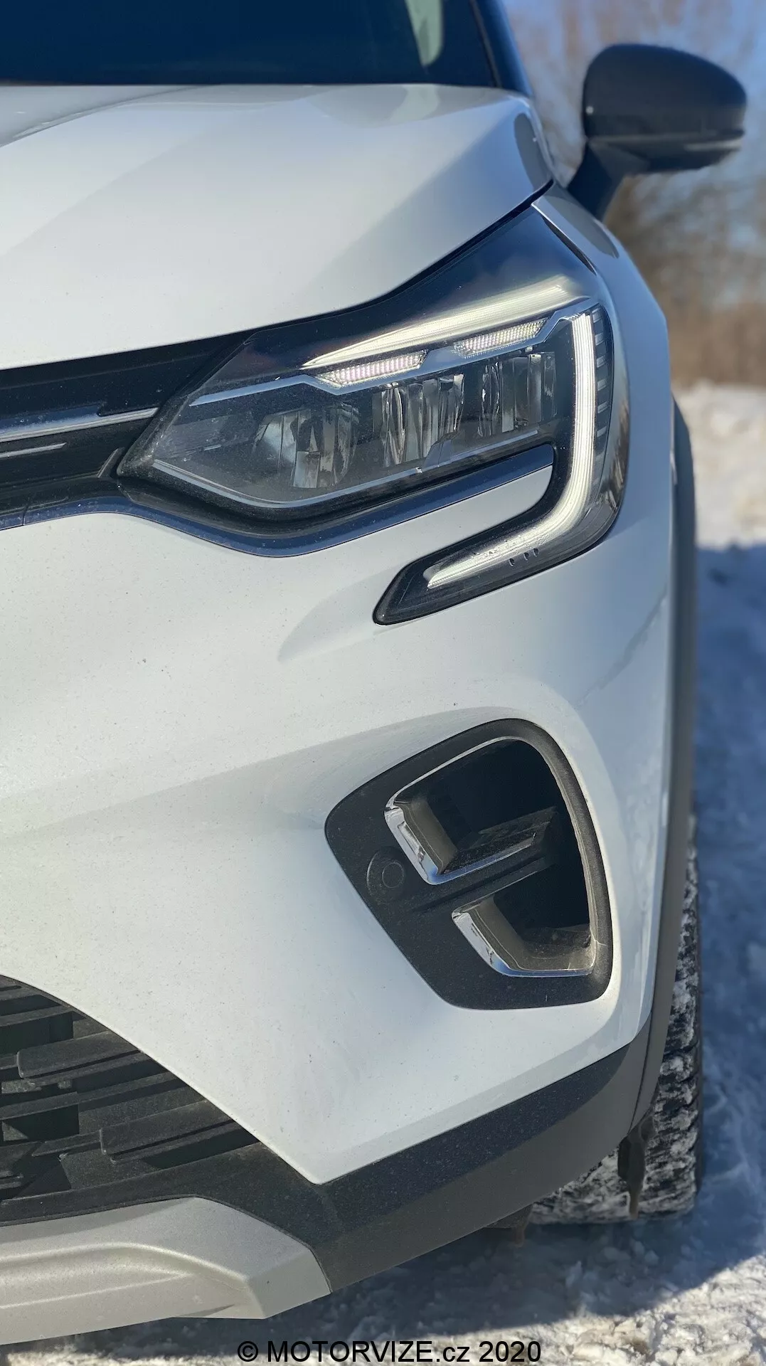 Primo piano del lato anteriore sinistro di una Renault Captur 2020 bianca, focalizzato sul fanale a LED moderno, il paraurti anteriore geometrico e gli elementi della griglia, catturato da un angolo frontale basso con neve sullo sfondo
