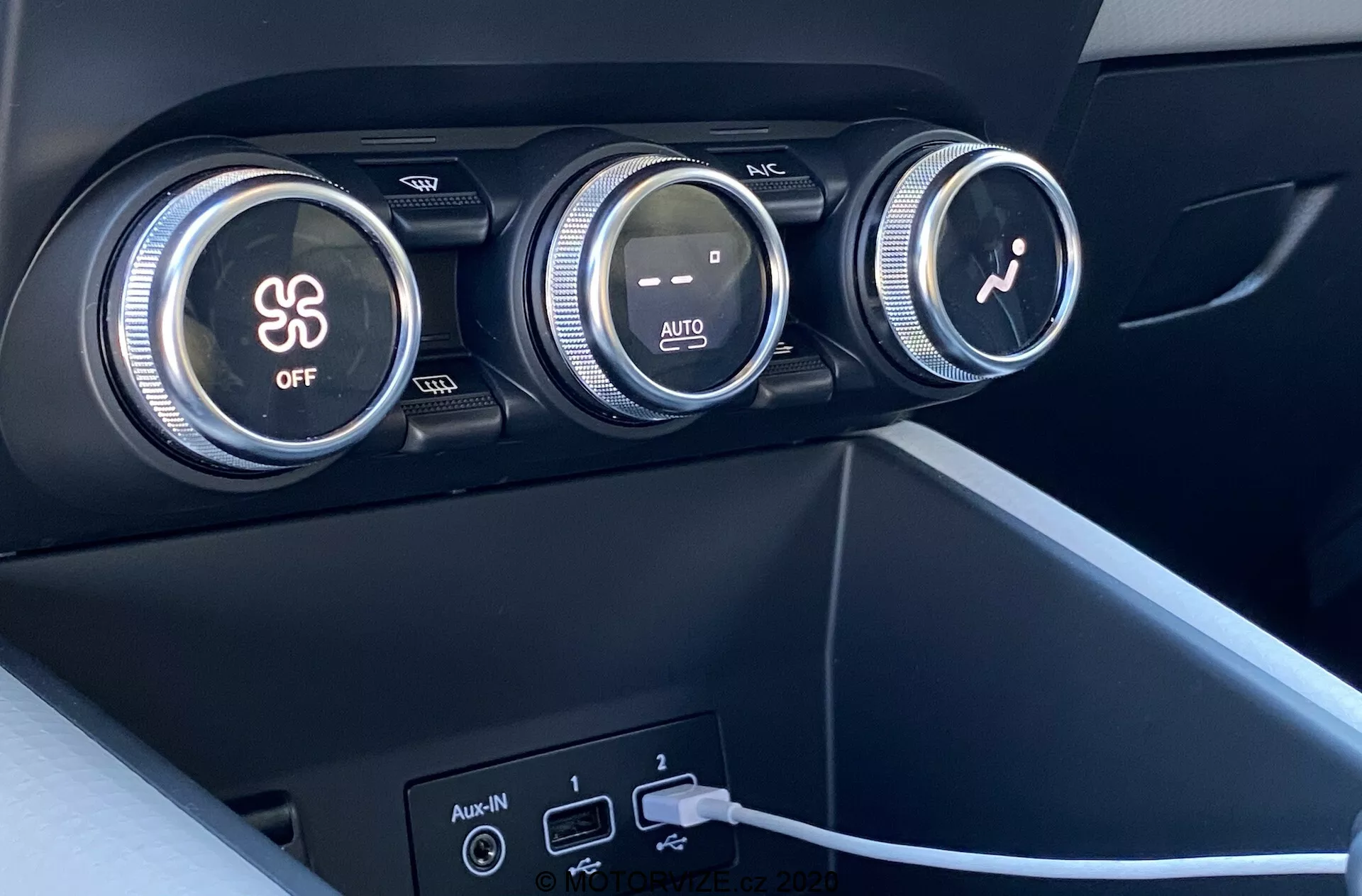 Primo piano degli interni della Renault Captur 2020 con particolare attenzione al sistema di climatizzazione. L'immagine mostra tre grandi manopole sul cruscotto. La prima manopola controlla il condizionatore d'aria, indicato da un simbolo di fiocco di neve e un pulsante 'OFF'. La manopola centrale regola la temperatura con gradienti di colore rosso e blu e un'indicazione 'AUTO'. La terza manopola con un'icona del ventilatore regola la velocità della ventola. Sotto le manopole ci sono due porte USB e un ingresso ausiliario, con un cavo collegato a una delle porte USB. I materiali sono di plastica scura con finitura opaca o sintetici e di design moderno e minimalista.