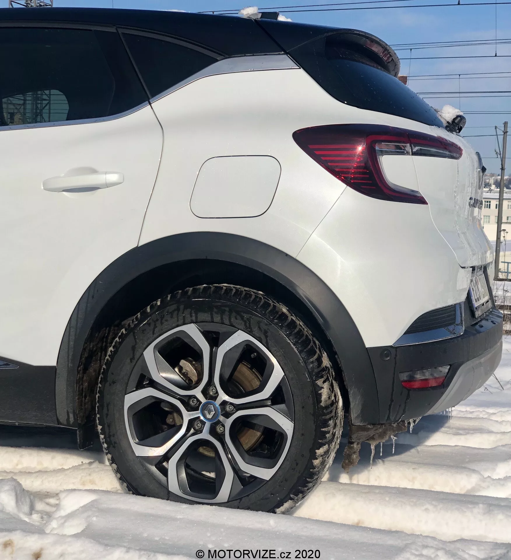 Primo piano dettagliato di una Renault Captur 2020 bianca, che mostra il lato passeggero posteriore da un'angolazione bassa. Si evidenziano la ruota posteriore con un design in lega multirazza, sporca di fango e neve, suggerendo condizioni invernali. L'immagine cattura il parafango posteriore pulito sopra l'arco della ruota, la moderna luce posteriore con sezioni a LED rosse e trasparenti, e il robusto rivestimento di plastica dietro la ruota. La finestra inclinata sul portellone posteriore contribuisce alla forma aerodinamica del veicolo.