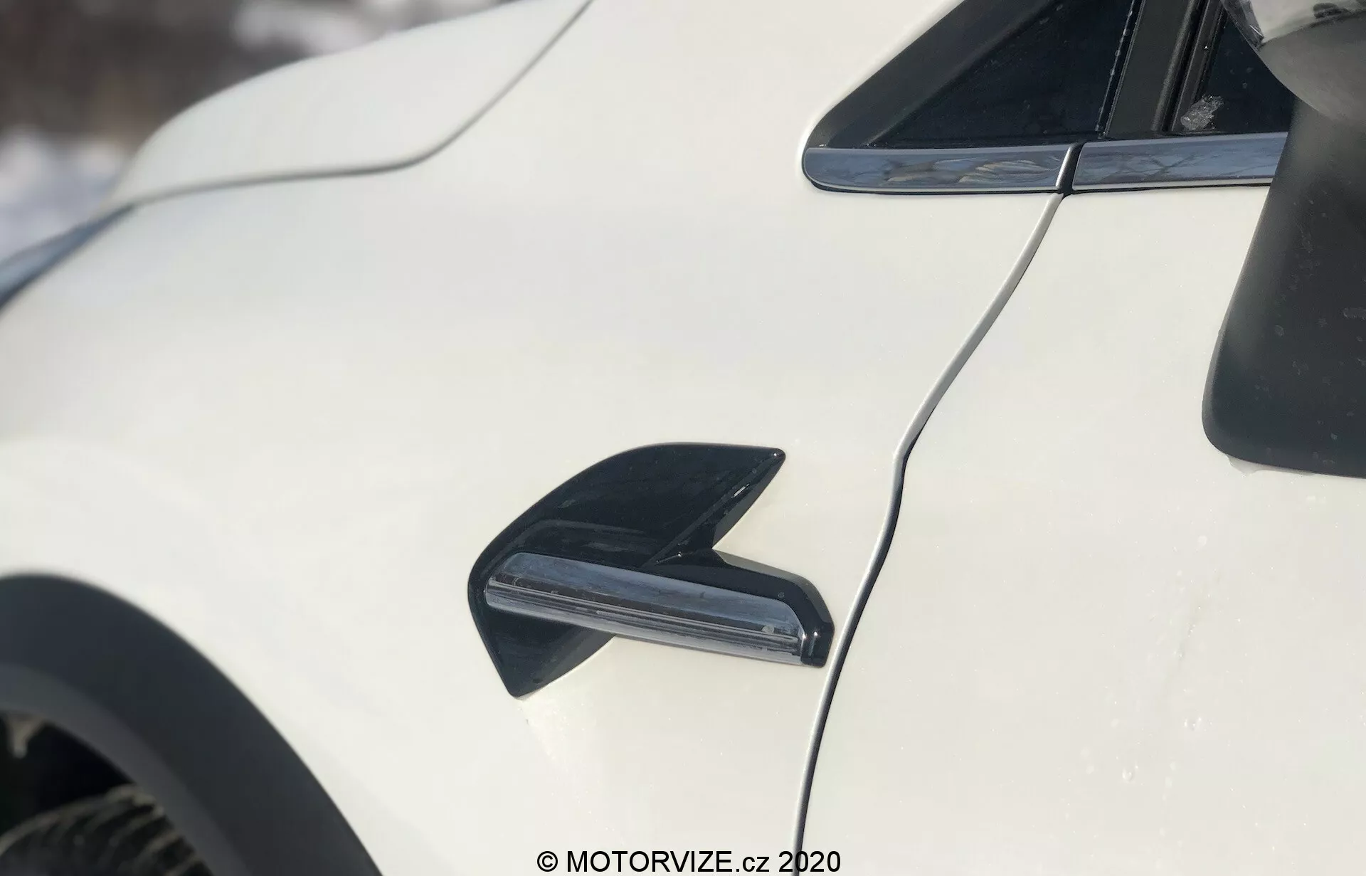 Primo piano del lato esterno della Renault Captur 2020 bianca. L'immagine presenta un'antenna nera a forma di pinna di squalo in alto a sinistra, progettata per la ricezione radio e l'aerodinamica. Sotto l'antenna, al centro, è evidenziata una presa d'aria nera, probabilmente per il raffreddamento dei freni o il miglioramento del flusso d'aria. Il profilo aerodinamico del tetto dell'auto si curva verso il retro, con la vernice bianca lucida che riflette la luce. Lo sfondo è leggermente sfocato, indicando un ambiente esterno innevato.