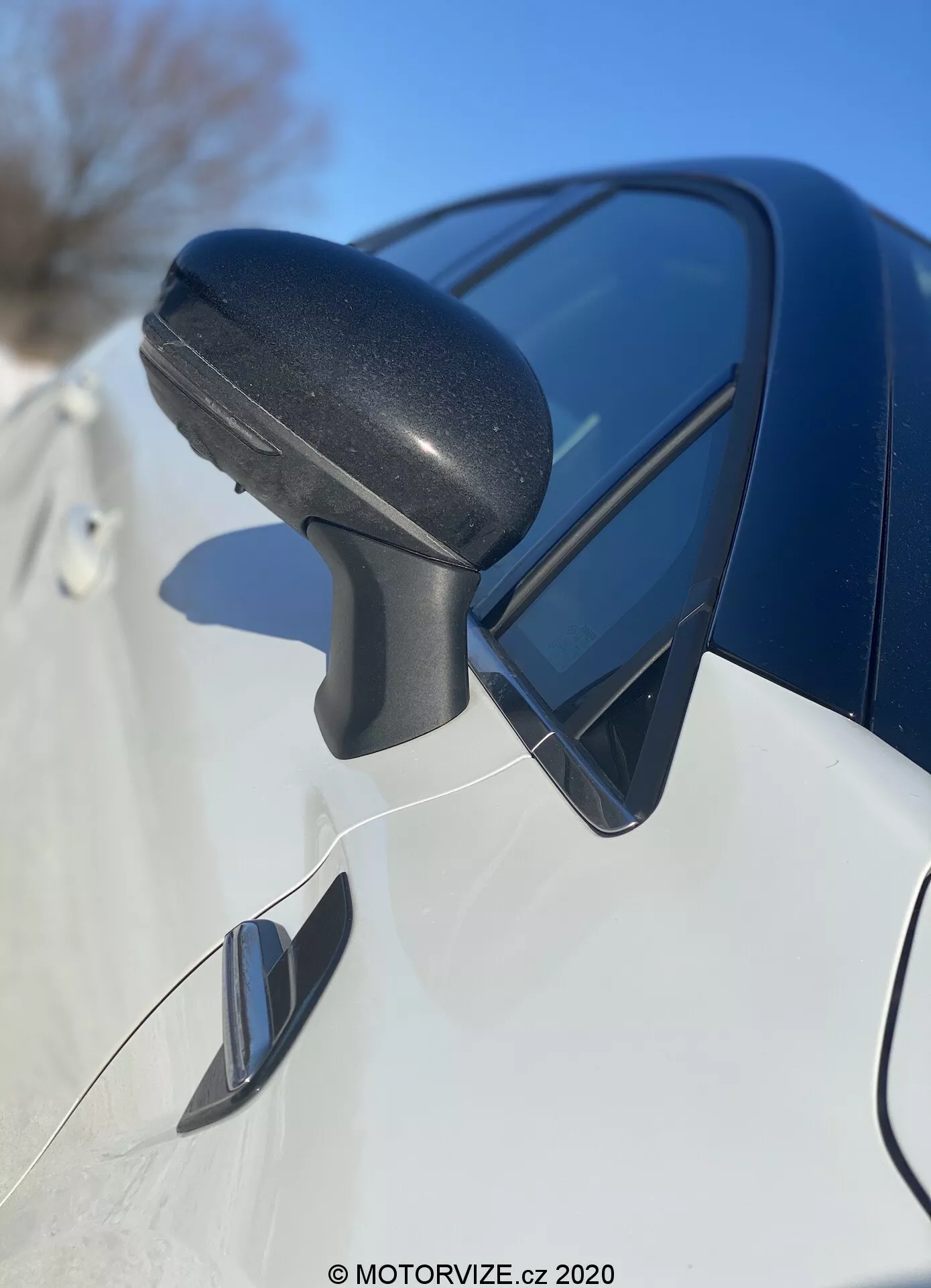 Vista ravvicinata dell'assemblea dello specchietto laterale della Renault Captur 2020 dalla parte anteriore a quella posteriore, con messa a fuoco sulla scocca dello specchio in plastica nera texturizzata con interruttore di segnalazione integrato e riflettente il cielo blu. Il pannello carrozzeria scuro e lucido sottostante e il possibile specchietto angolare indicano un design moderno e caratteristiche avanzate.