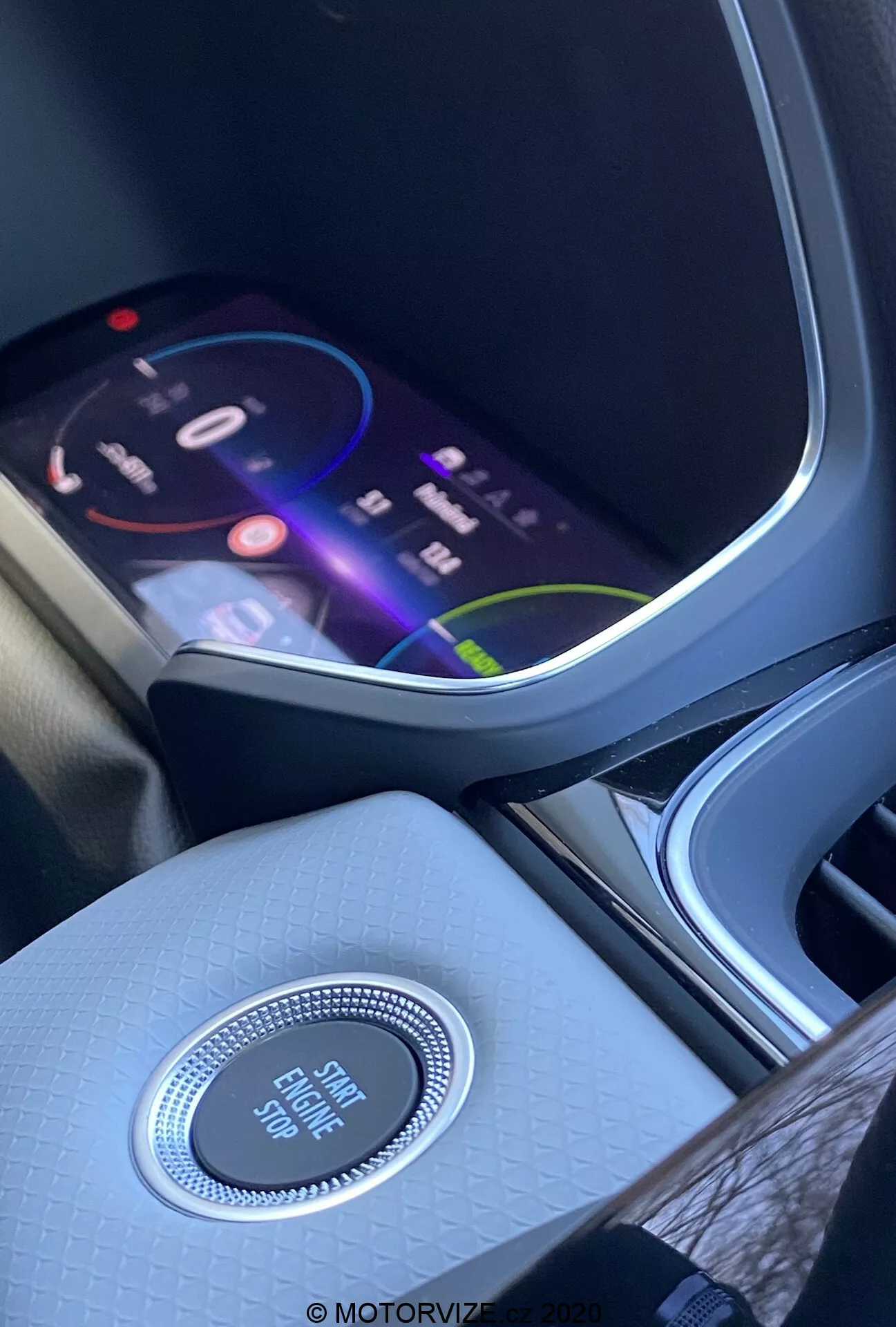 Interni della Renault Captur 2020 focus sulla console centrale e il cruscotto dalla prospettiva del sedile anteriore. Lo schermo di infotainment vivace mostra un'interfaccia colorata che suggerisce funzioni multimediali avanzate, tra cui navigazione, riproduzione di media e opzioni di connettività. Sotto lo schermo, un pomello di controllo circolare con finitura cromata etichettato 'Engine Start Stop' evidenzia il sistema di avvio senza chiave. La console centrale presenta una finitura testurizzata di alta qualità, migliorando l'ambiente interno. A sinistra, è visibile parzialmente un portabicchieri o un vano portaoggetti.