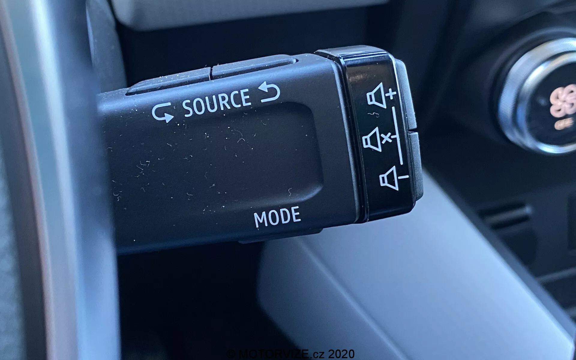 Primo piano dei comandi audio montati sul volante di una Renault Captur 2020, concentrandosi sulla leva di controllo sul lato sinistro con pulsanti per cambiare la sorgente (SOURCE), regolare il volume, saltare tracce e selezionare la modalità (MODE). La fotografia cattura i dettagli dei pulsanti in plastica con finitura opaca e del loro design ergonomico, con parte della colonna dello sterzo e dell'interno sfocati sullo sfondo, evidenziando la funzionalità e la praticità per il conducente.
