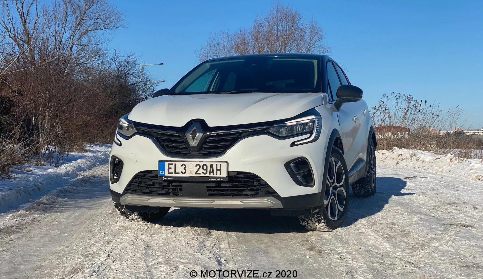 Vista frontale di una Renault Captur bianca del 2020, fotografata con un'angolazione che favorisce leggermente il lato sinistro. Il veicolo moderno mostra un distintivo design della griglia frontale Renault con un ampio logo centrale, linee angolari affilate e contorni scolpiti. La griglia frontale, i fari a LED e le luci diurne angolari sono chiaramente visibili. Il paraurti sportivo presenta prese d'aria laterali prominenti e una sezione centrale probabilmente per il raffreddamento. Il cofano ha linee rialzate per un aspetto muscoloso. Una parte delle ruote anteriori e dei cerchi in lega a più razze è visibile sulla superficie innevata, indicando condizioni invernali. La targa europea e la vernice lucida che riflette la luce solare suggeriscono una giornata serena e soleggiata. È possibile prevedere sistemi avanzati di assistenza alla guida, illuminazione a LED e propulsione ibrida/elettrica dall'iniziale 'E' sulla targa.