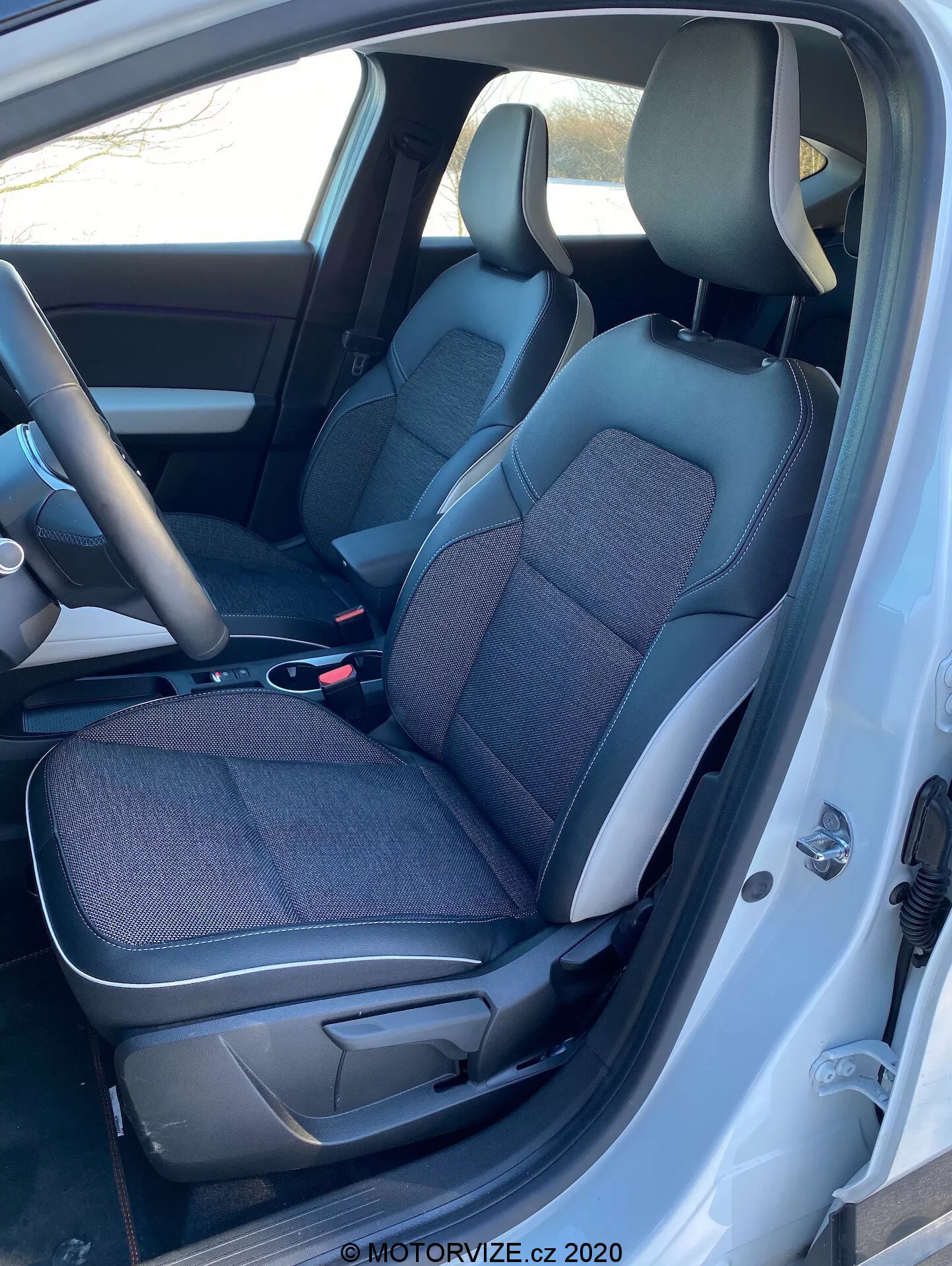 Vista interna del Renault Captur 2020 dall'interno del lato sinistro con la porta del conducente aperta, mostrando sedili in tessuto e pelle neri e grigi, consolle centrale, volante e parte del cruscotto.