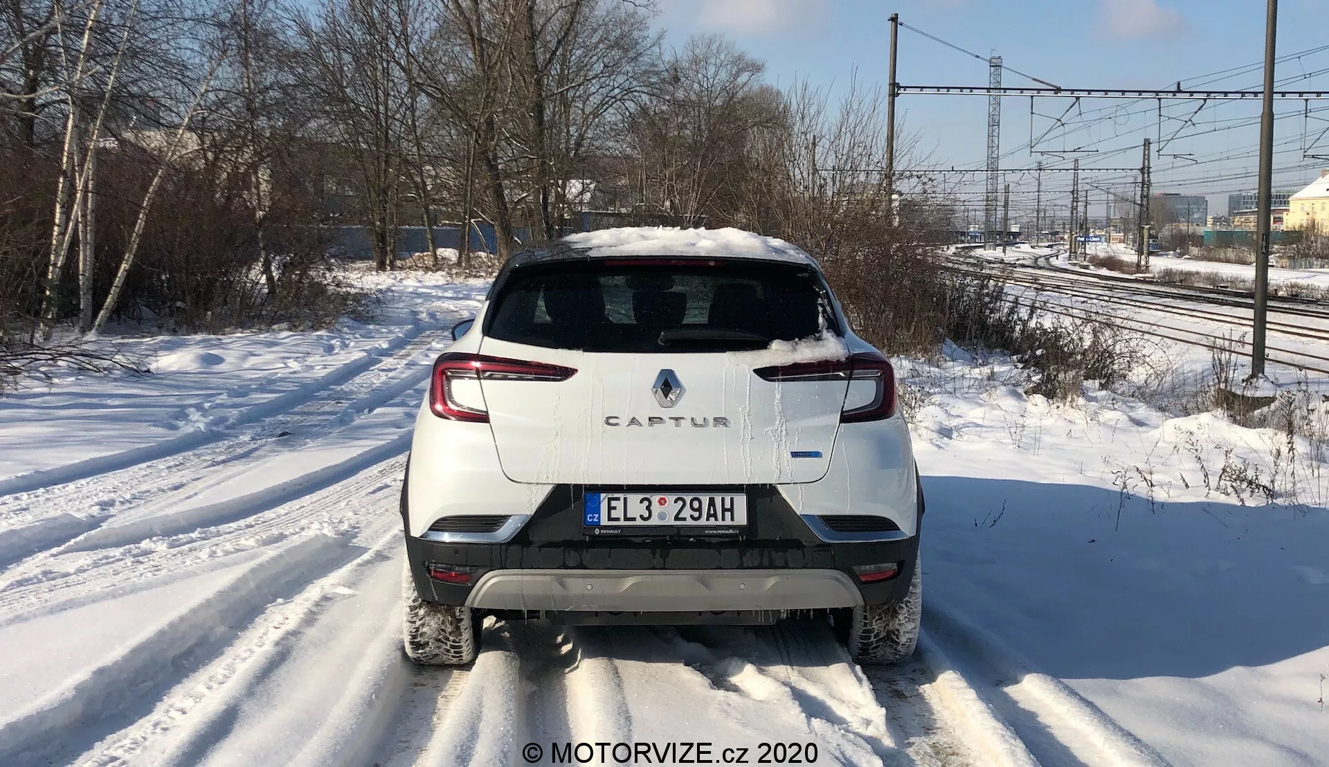 Vista posteriore centrata di una Renault Captur 2020 su una strada innevata. L'immagine mette in evidenza il design posteriore distintivo, inclusi lo schema di colore bicolore con carrozzeria bianca e tetto a contrasto, l'illuminazione posteriore a LED moderna, la targa in stile europeo, il tergicristallo posteriore e un unico tubo di scarico sul lato destro. La foto cattura linee elettriche e alberi sullo sfondo alla luce del giorno, suggerendo un ambiente suburbano o semirurale.