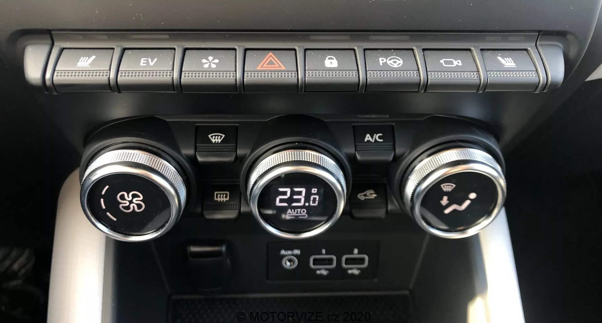 Vista ravvicinata del pannello di controllo del clima del Renault Captur 2020 all'interno del veicolo, che mostra tre manopole e diversi pulsanti. La manopola sinistra controlla la velocità della ventola, la manopola centrale con display digitale è impostata a 23.0°C per la temperatura e la manopola destra dirige il flusso d'aria all'interno dell'abitacolo. Sopra le manopole, una fila di pulsanti include ricircolo dell'aria, sbrinamento del parabrezza, sbrinamento del lunotto posteriore, sincronizzazione della temperatura, interruttore modalità Auto e pulsante EV che indica capacità di veicoli elettrici. Sotto la manopola centrale ci sono due porte USB e una presa di corrente da 12V, evidenziando la connettività moderna e le opzioni di ricarica.