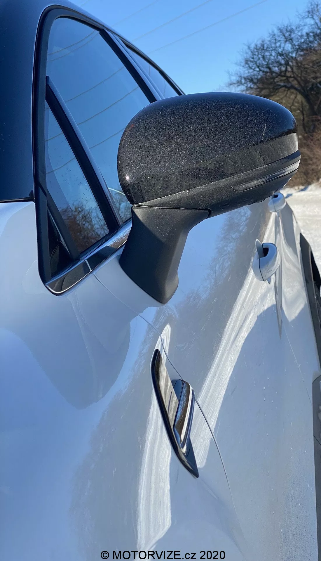 Vista dettagliata da vicino dello specchietto laterale, della porta e del quarto pannello anteriore di una Renault Captur 2020 bianca. La foto, scattata dal lato appena davanti allo specchietto laterale con un angolo verso l'alto, mette in evidenza il design moderno ed efficiente aerodinamico dello specchietto laterale con una sezione inferiore nera testurizzata per durata. Il supporto aerodinamico si integra perfettamente nel telaio della portiera. La maniglia della porta incassata e la linea caratteristica che la attraversa suggeriscono un design sportivo e snello. Una parte del faro e il riflesso del cielo chiaro nel finestrino esaltano questa presentazione dinamica.