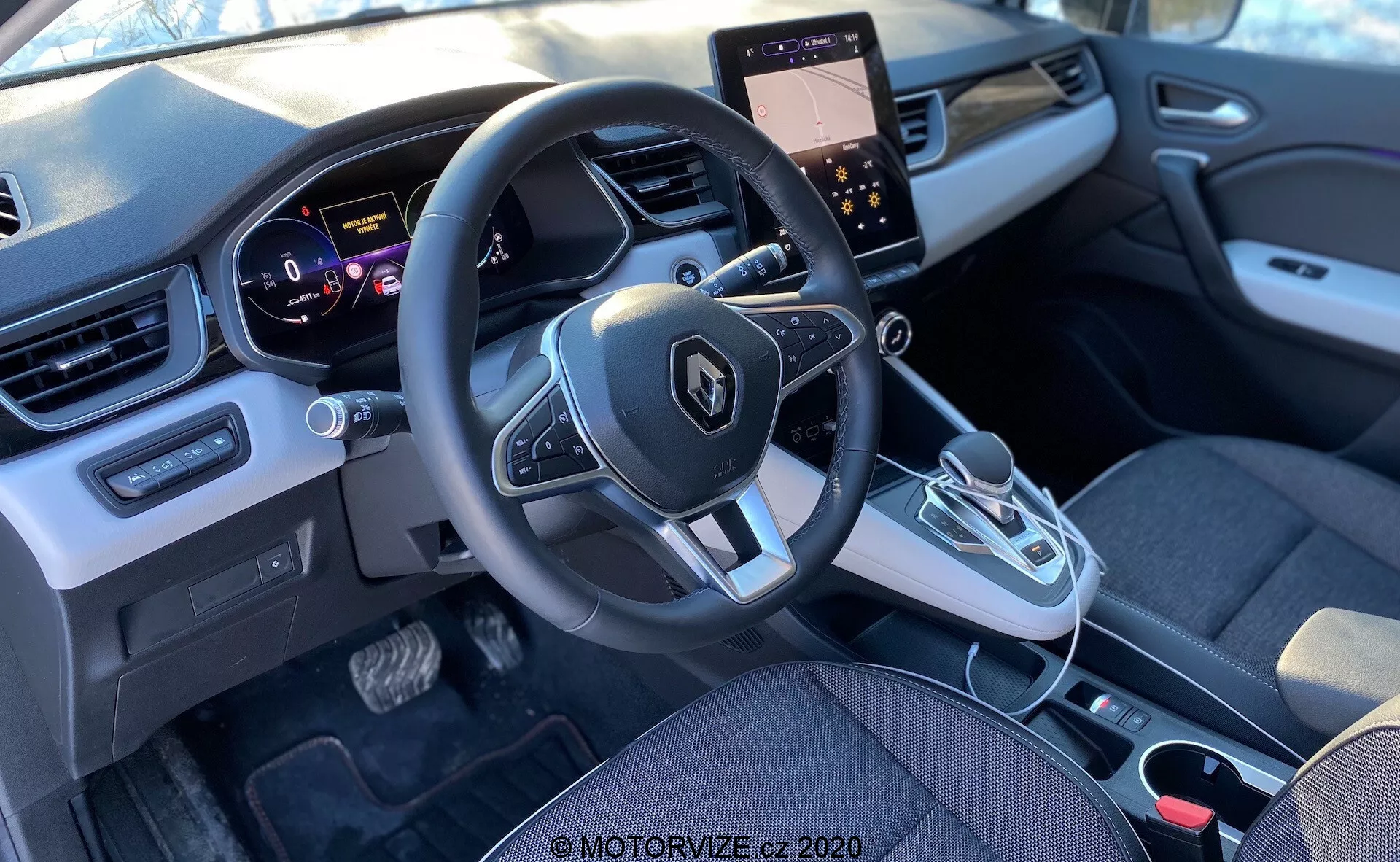 Vista interna di una Renault Captur 2020 dal sedile posteriore, con focus sull'area della cabina anteriore che comprende il cruscotto, il volante con controlli integrati, il quadro strumenti, la console centrale con display touchscreen e comandi per il clima, leva del cambio automatico e un design moderno che combina tessuto e plastica.