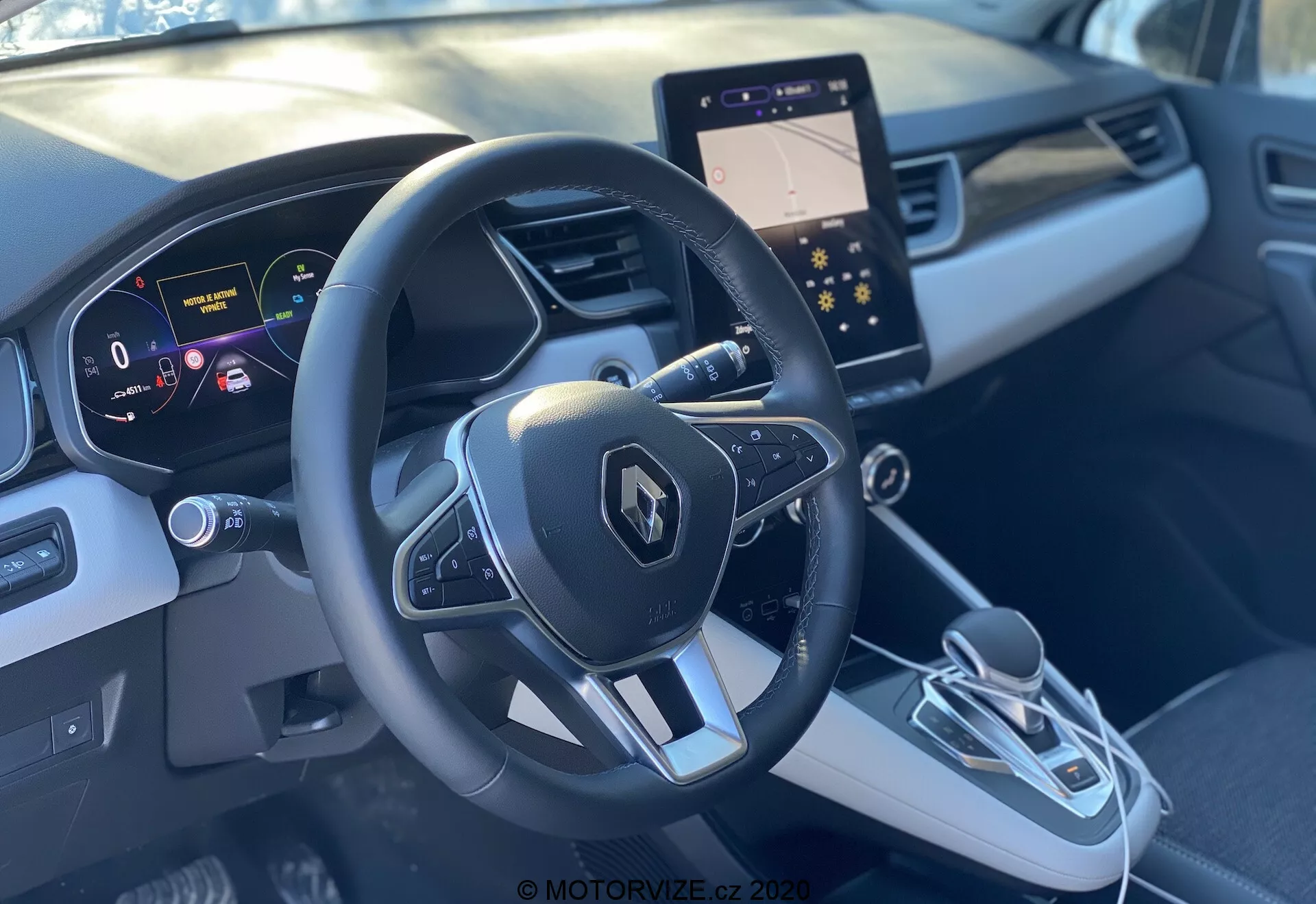 Vista degli interni della Renault Captur 2020 dal sedile del passeggero anteriore, che mostra il volante sportivo a tre razze con emblema Renault e pulsanti multifunzione, quadro strumenti digitale con display ad alta risoluzione che mostra la velocità e gli aiuti alla navigazione, e la console centrale elegante. La console centrale presenta un moderno schermo di infotainment, selettore del cambio elettronico e pulsante del freno di stazionamento elettronico. L'interno bicolore grigio con design sagomato e rifiniture offre una sensazione di lusso.