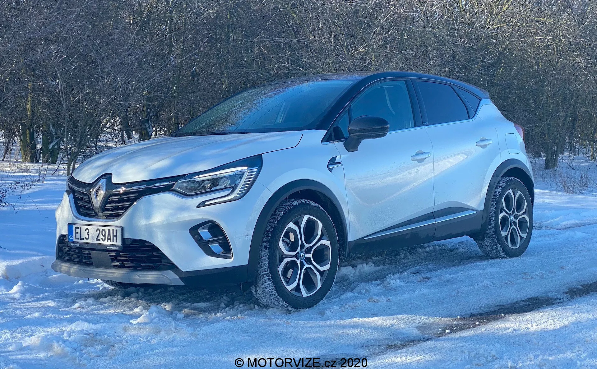 Vista dal basso di un SUV Renault Captur 2020 su una superficie innevata, che mostra il suo profilo del quarto anteriore. La luce del giorno brillante e il cielo limpido mettono in risalto gli elementi di design moderno come la griglia frontale prominente con badge, le luci diurne a LED, le complesse ruote in lega bicolore, la carrozzeria scolpita con passaruota pronunciati, gli specchietti in tinta con la carrozzeria con indicatori di direzione integrati, i finestrini oscurati per la privacy e le maniglie delle porte a filo. La foto enfatizza la costruzione premium del veicolo e le sue potenziali capacità di guida invernale.