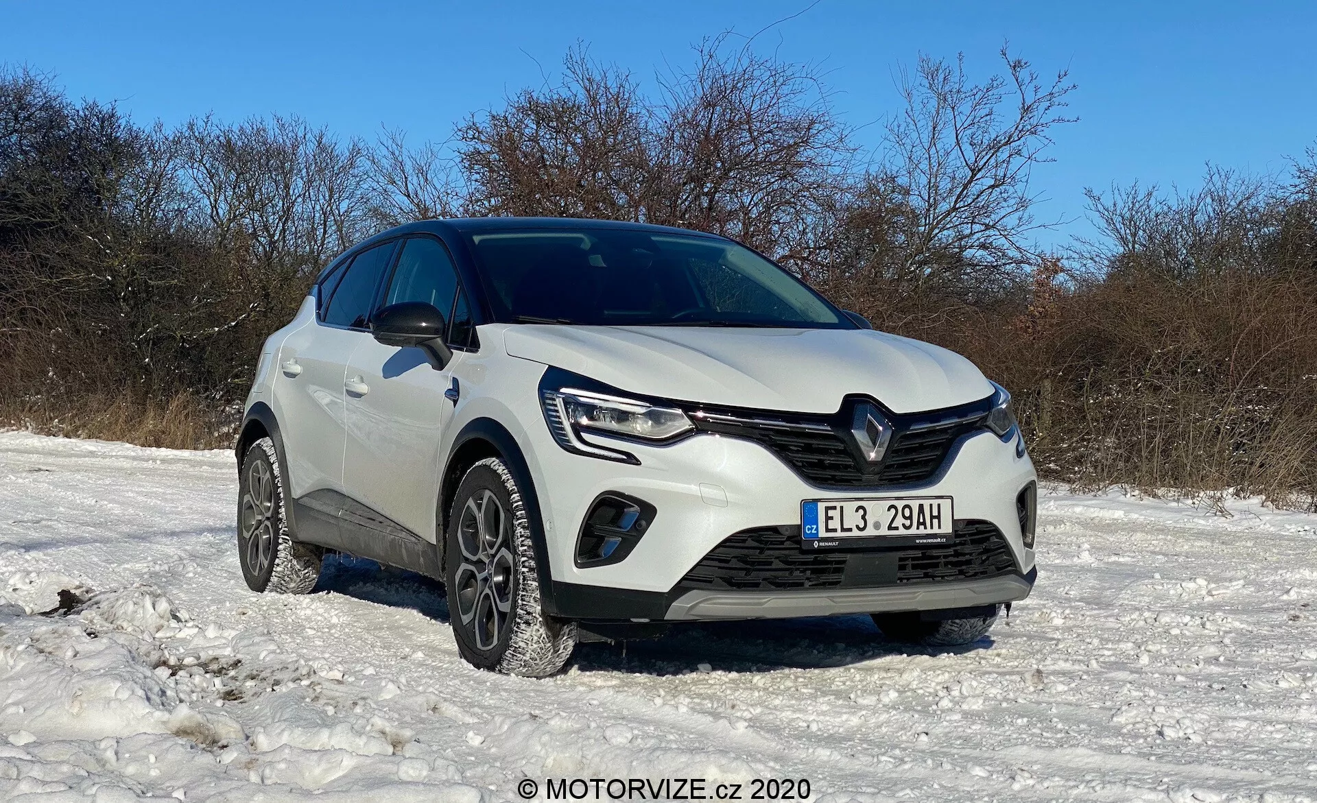 Renault Captur 2020 bianco parcheggiato su un terreno coperto di neve sotto un cielo terso, mostrato da un angolo frontale a tre quarti basso, con griglia anteriore, fari con luci diurne a LED, fendinebbia, paraurti anteriore, pneumatici invernali, logo Renault distintivo, linee scolpite sul cofano, altezza elevata, possibili sensori di parcheggio assistito, specchietti laterali e condizione generale pulita e ben tenuta, con filigrana MOTORVIZE.cz © 2020.