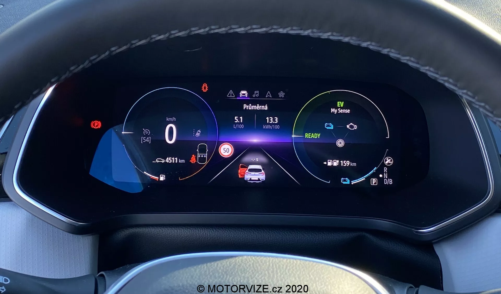 Vista ravvicinata del quadro strumenti digitale dell'auto moderna Renault Captur 2020 dalla prospettiva del conducente. Il display completamente digitale ad alta risoluzione a colori, inserito in un cruscotto testurizzato con cuciture visibili, mostra un tachimetro a 0 km/h con un limite di velocità di 50 km/h, un grafico animato centrale dell'auto con avvisi di sicurezza e indicatori di batteria, un tachimetro con icona della modalità ecologica 'My Sense', contachilometri, temperatura esterna, indicatore della marcia in 'Park' e icone per assistenza al mantenimento della corsia, abbaglianti automatici e controllo di crociera adattivo, tutto in un layout ben organizzato che evidenzia la tecnologia automobilistica avanzata e il design degli interni.