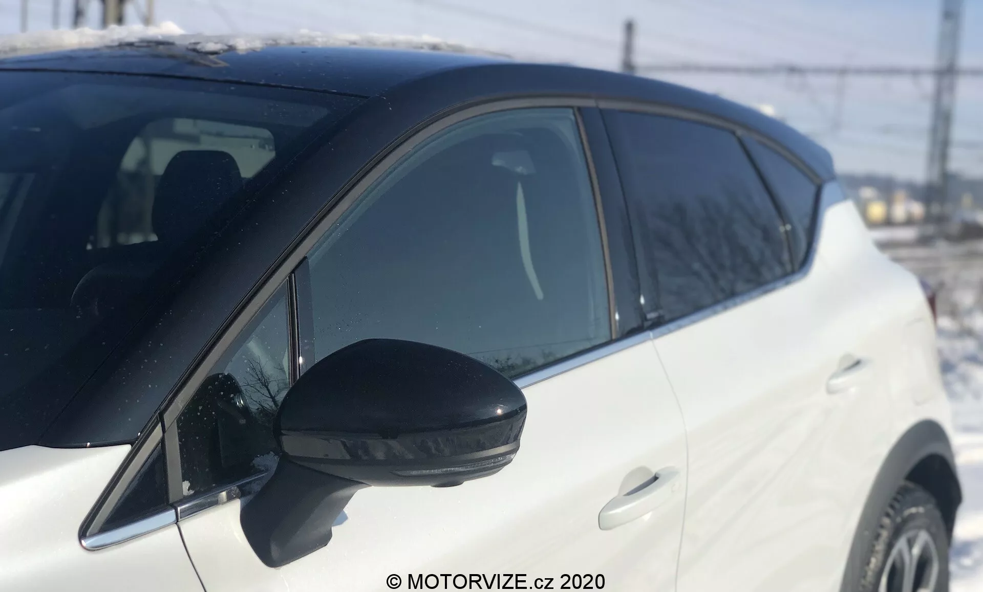 Vista laterale ravvicinata di una Renault Captur 2020 bianca, con messa a fuoco dallo specchietto laterale fino a prima della ruota anteriore. Specchietto laterale nero prominente con indicatore di direzione integrato, fotografato alla luce del giorno con luce naturale. Angolo leggermente basso che enfatizza la sezione centrale, profondità di campo ridotta. Sfondo sfocato con elementi discernibili come recinzione, linee elettriche e una struttura, che indicano un ambiente esterno. Vernice bianca lucida del veicolo e maniglie delle porte abbinate al colore, mostrando un design moderno ben mantenuto. Linea di finestrino ascendente verso la parte posteriore che indica uno stile dinamico.