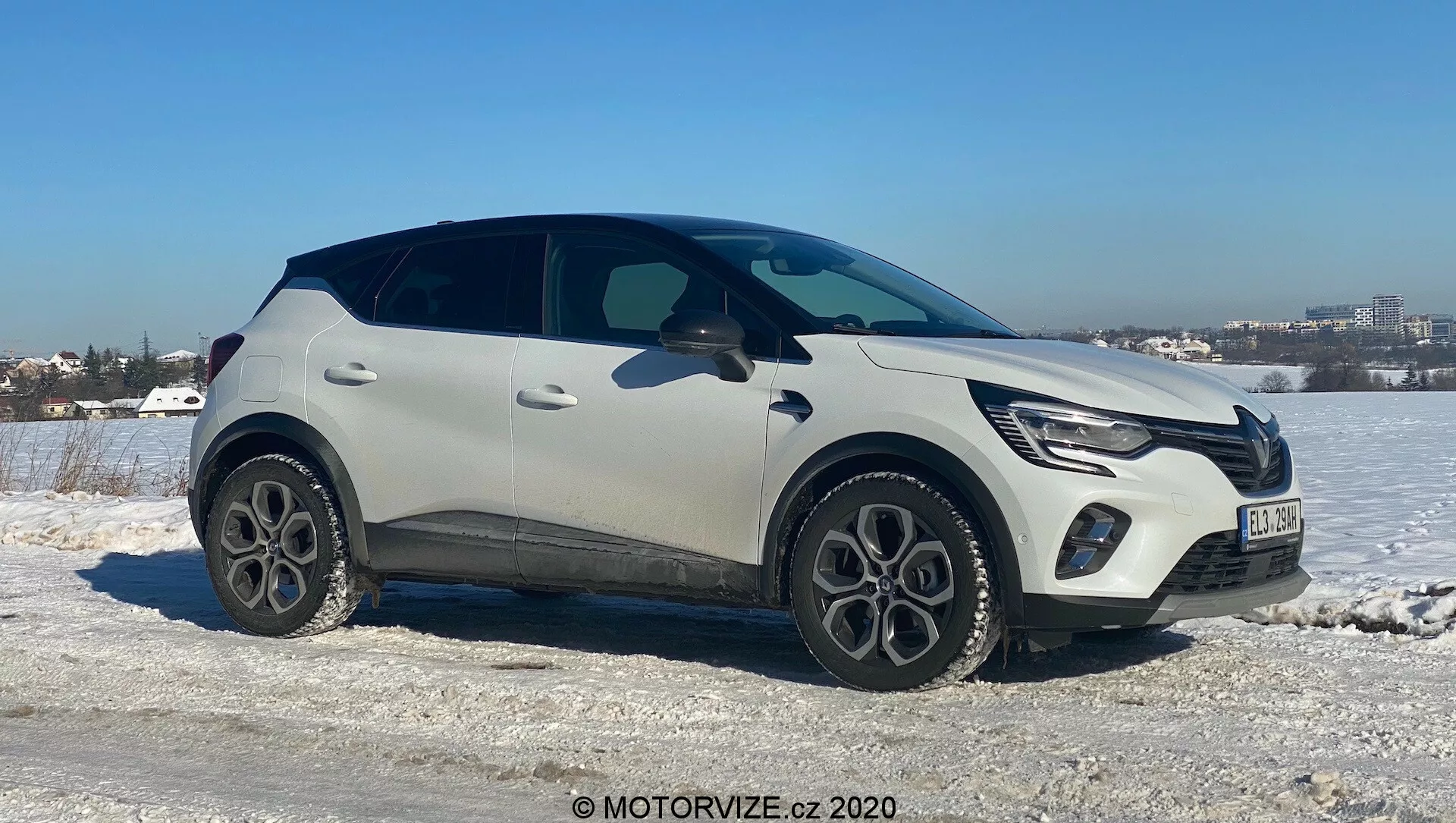 Renault Captur 2020 moderno crossover SUV posizionato su un terreno ghiacciato e innevato con un cielo limpido sullo sfondo. Il lato passeggeri è rivolto verso la fotocamera con un angolo leggermente inclinato, mostrando la parte anteriore, laterale e parzialmente superiore del veicolo. Caratteristiche includono luci diurne a LED, griglia frontale integrata con emblema del produttore, verniciatura bicolore chiara e nera, cerchi in lega a cinque razze, modanature protettive in plastica, maniglie delle porte eleganti e incassate e una linea dei finestrini affusolata con spoiler posteriore. Suggerisce tecnologia avanzata come l'illuminazione a LED e l'ingresso senza chiave, ambientati in un paesaggio in condizioni climatiche fredde vicino a una zona urbana.
