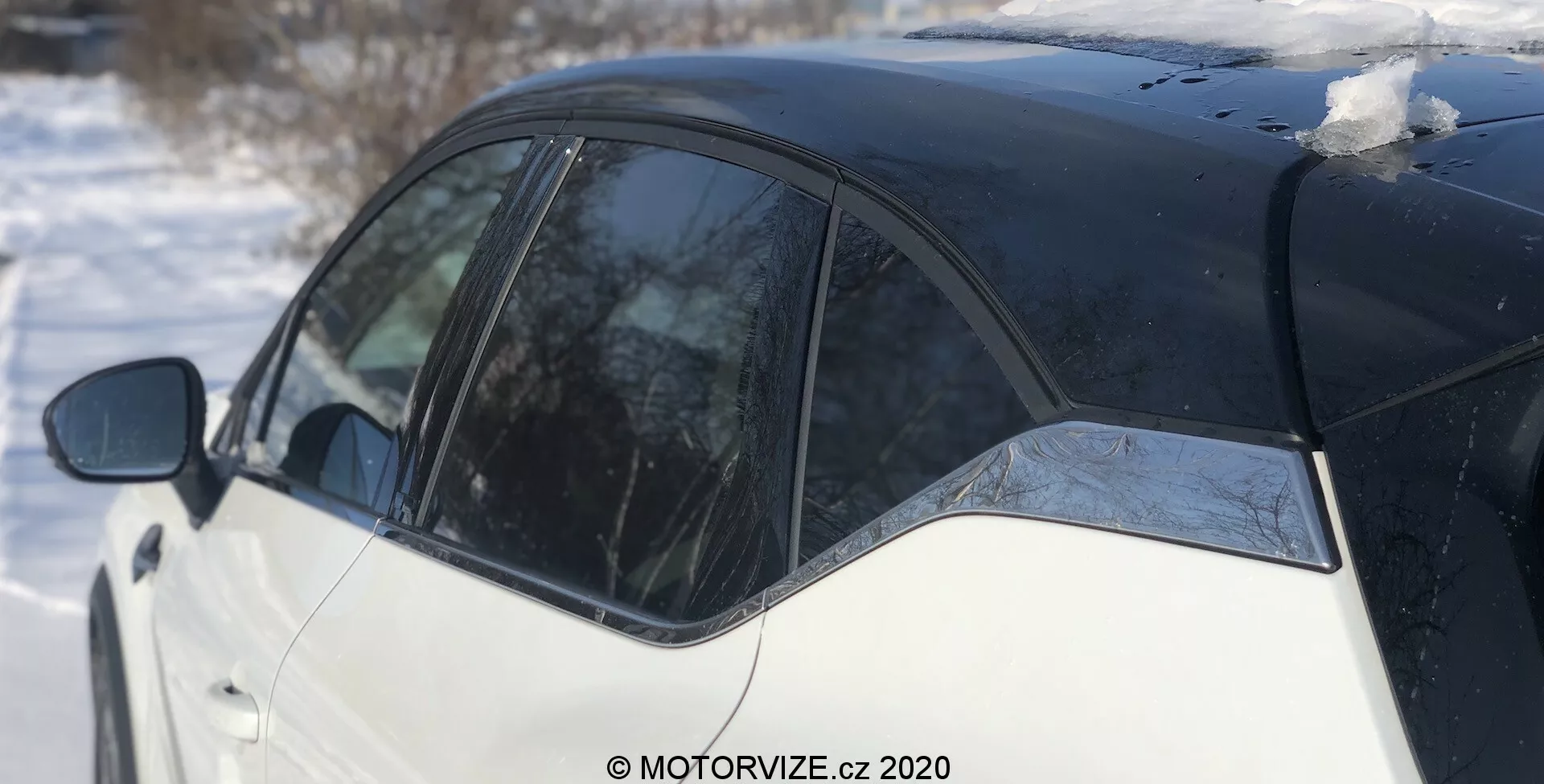Vista laterale della Renault Captur 2020 in un'ambientazione invernale, con evidenziato uno schema di vernice bicolore bianco e nero. L'immagine si concentra sul montante B, su parte della porta posteriore e sul pannello posteriore, catturato da una prospettiva leggermente arretrata. Macchie di neve sul tetto e ghiaccio che si scioglie sul finestrino riflettono una giornata soleggiata.