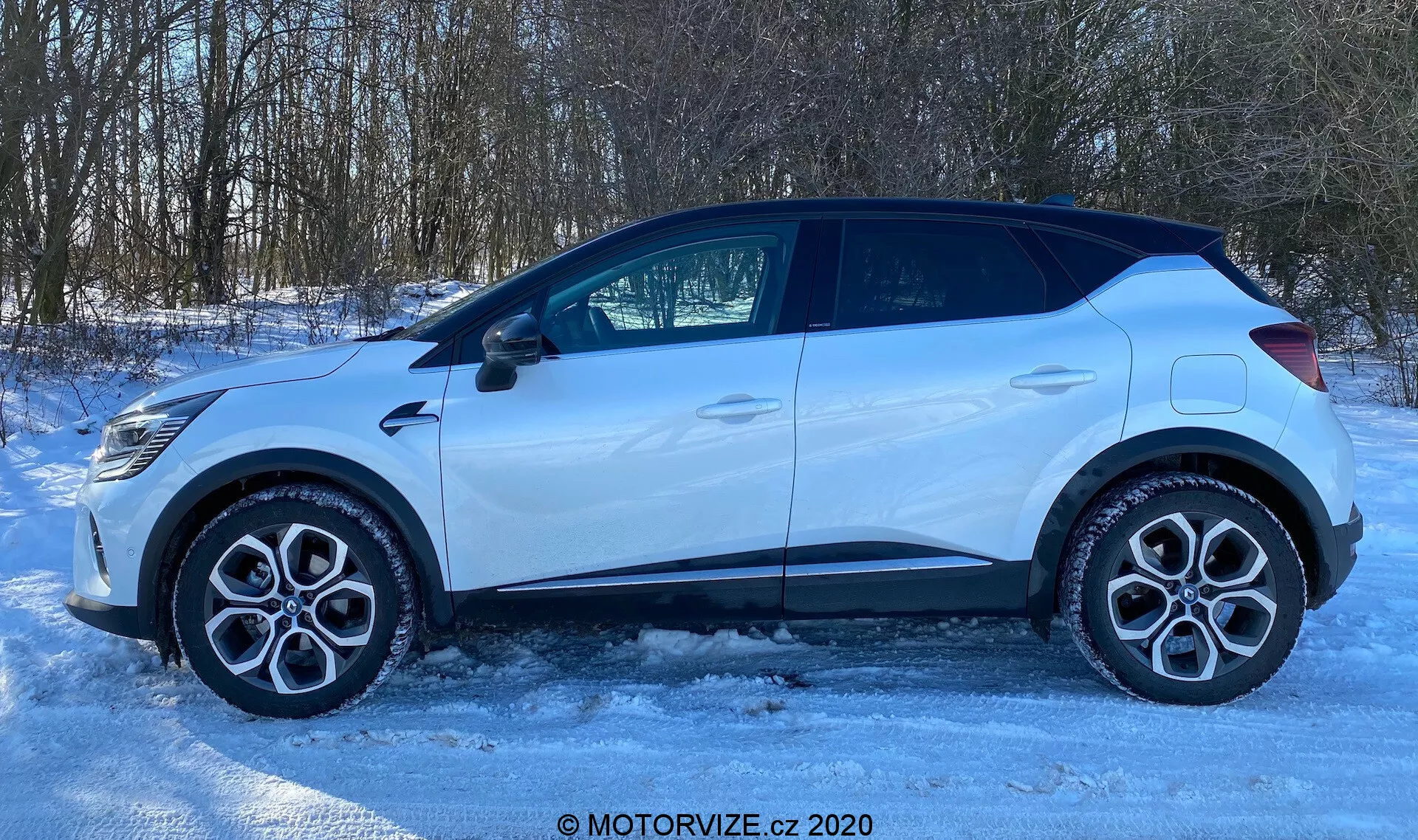 Vista laterale di una Renault Captur 2020 parcheggiata su una superficie ghiacciata o innevata. Il profilo dell'auto mostra cerchi in lega, pneumatici stradali standard, quattro porte, finestre laterali, inclusa una piccola finestra posteriore, e uno specchietto retrovisore laterale con indicatore di direzione integrato. La linea del tetto è leggermente inclinata verso la parte posteriore, con rivestimenti in plastica intorno ai passaruota e alla parte inferiore delle portiere, suggerendo un design pronto per fuoristrada. La scena ben illuminata, probabilmente dalla luce solare naturale, definisce la forma dell'auto contro un ambiente rurale o suburbano con alberi sullo sfondo. L'immagine ha una filigrana che riporta la scritta 'MOTORVIZE.cz 2020'.