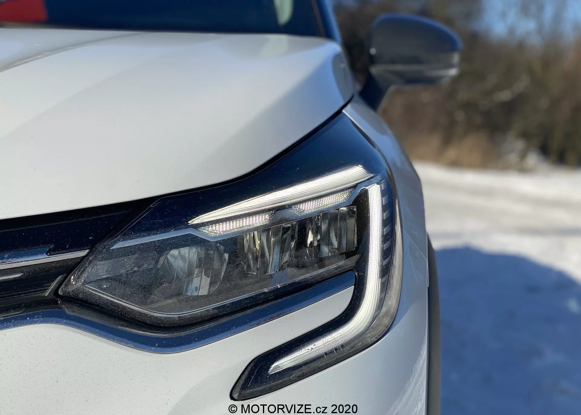 Vista ravvicinata dell'angolo anteriore sinistro di un'auto moderna, che si concentra sul faro a LED e sulla griglia, presa da un angolo basso vicino al livello del faro. Le luci diurne presentano un design curvo che abbraccia l'unità del faro. Fari adattivi o luci di svolta angolare visibili, con finitura della vernice metallica e indicatori di direzione integrati nello specchietto retrovisore esterno. L'illuminazione naturale brillante suggerisce un'ambientazione diurna all'aperto.