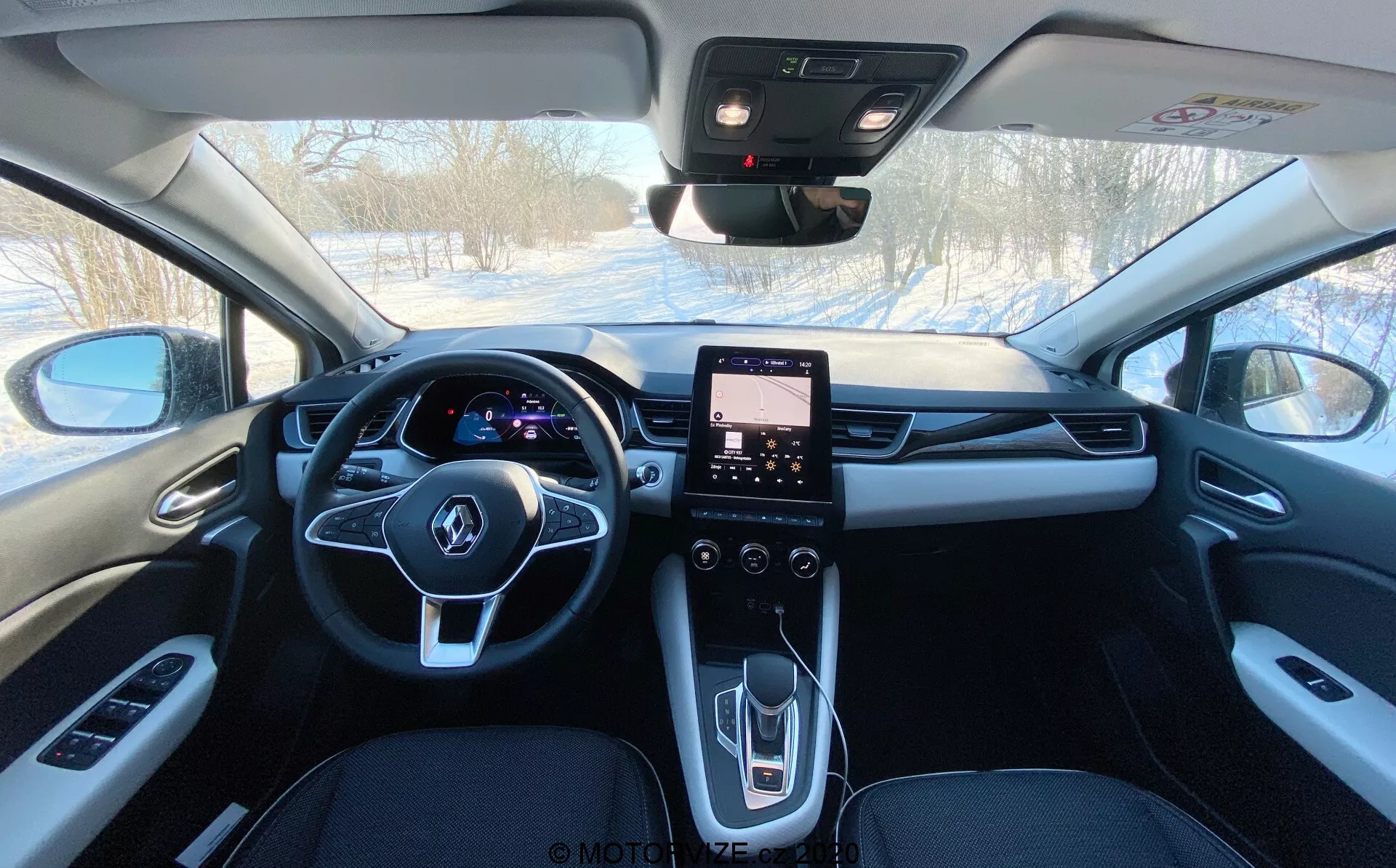Vista interna di una Renault Captur 2020 dall'area dei sedili posteriori verso il cruscotto, mostrando la tappezzeria di colore scuro, lo specchietto retrovisore centrale, il cruscotto digitale illuminato dietro al volante multifunzione, sistema di infotainment touchscreen nel quadro centrale e i controlli per i finestrini elettrici, specchietti laterali, controllo del clima, e probabilmente per il tettuccio apribile e il pulsante SOS di emergenza.