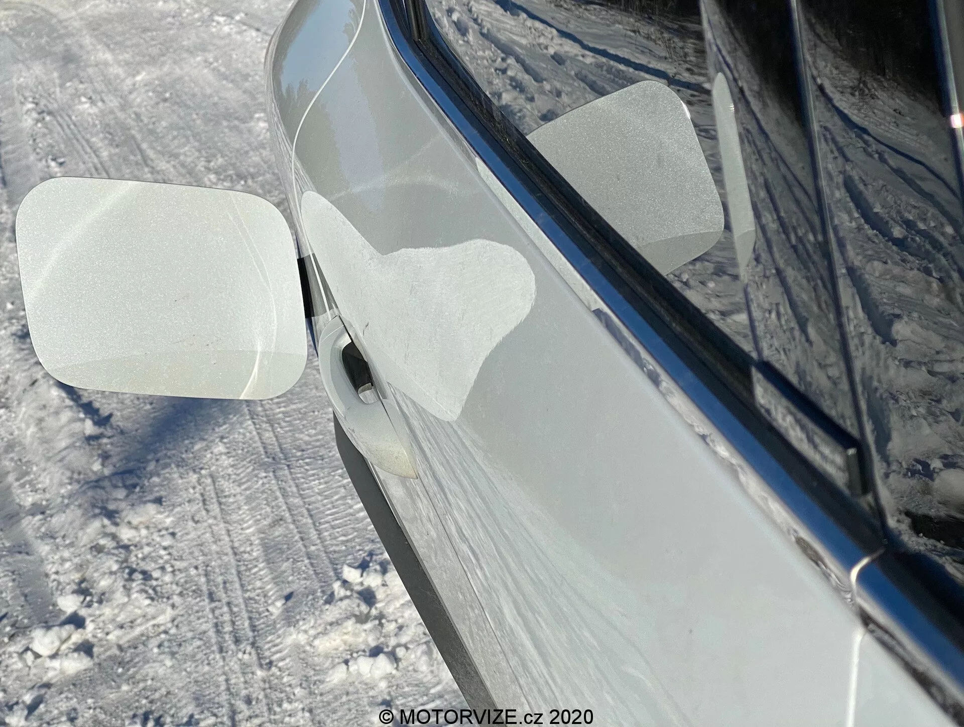 Vista laterale ravvicinata di una Renault Captur 2020 in un ambiente nevoso all'aperto. L'attenzione è rivolta allo specchietto retrovisore destro, ripiegato in avanti, indicando una possibile funzione di piegatura automatica o manuale. La carcassa dello specchietto ha due pellicole protettive trasparenti a forma di parallelogramma, probabilmente per protezione durante la consegna o il lavaggio. L'auto, di un colore scuro lucido come nero o blu scuro, appare ben mantenuta con riflessi che suggeriscono una superficie pulita e lucidata. Il terreno circostante e i lati dell'auto sono coperti di neve. È visibile anche una parte della maniglia della porta anteriore con un pulsante di accesso senza chiave o un sensore touch.