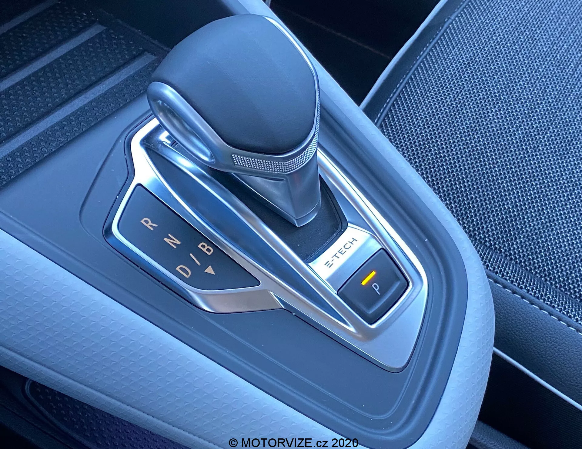 Immagine ravvicinata di una moderna leva del cambio automatico in una Renault Captur 2020, situata sulla console centrale. Il selettore del cambio elettronico è posizionato verticalmente ed è realizzato in pelle, plastica e accenti metallici. La foto, scattata da una prospettiva dall'alto leggermente angolata a destra, mette in evidenza le indicazioni delle marce (P, R, N, D, B) illuminate da luci a LED. A destra è visibile un pulsante con la scritta 'P', probabilmente per il freno di stazionamento elettronico. L'area intorno alla leva del cambio presenta plastica testurizzata di alta qualità e accenti metallici, riflettendo la tecnologia avanzata e il design contemporaneo degli interni del veicolo.