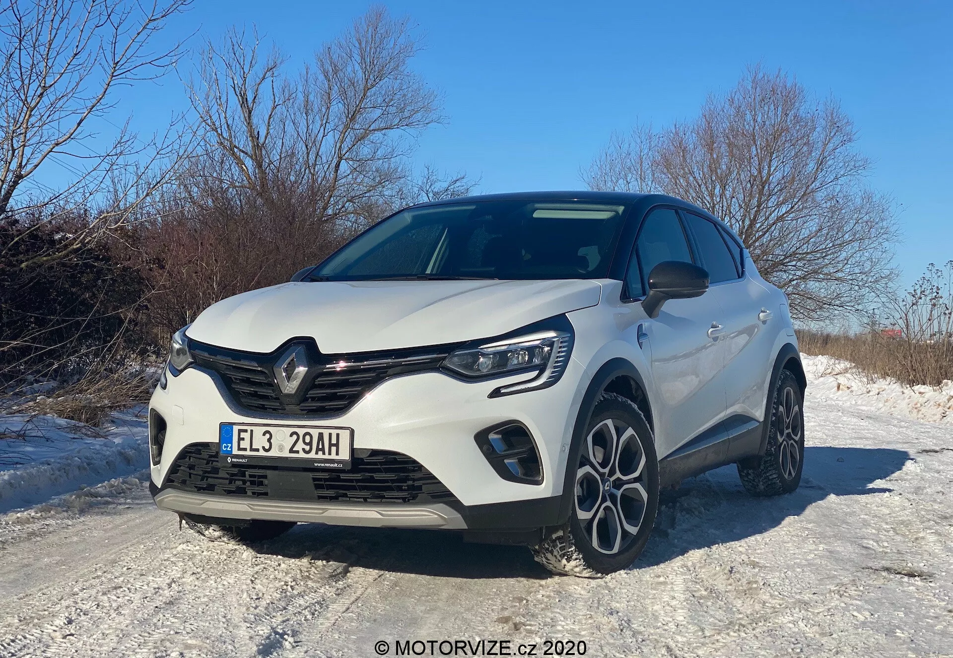 Veicolo moderno Renault Captur 2020 catturato da un angolo frontale basso a tre quarti, che mostra la griglia anteriore, il cofano, i fari a LED, il paraurti anteriore con luci fendinebbia integrate e un profilo laterale parziale. Gli elementi di design includono una griglia prominente con il logo del produttore, linee scolpite sul cofano e cerchi in lega a più raggi, il tutto sotto un cielo chiaro diurno, riflettendo un'estetica sportiva e aerodinamica, possibilmente evidenziando una versione potenziata o un pacchetto sportivo. L'auto è situata su una strada innevata, suggerendo caratteristiche adatte alla guida invernale.