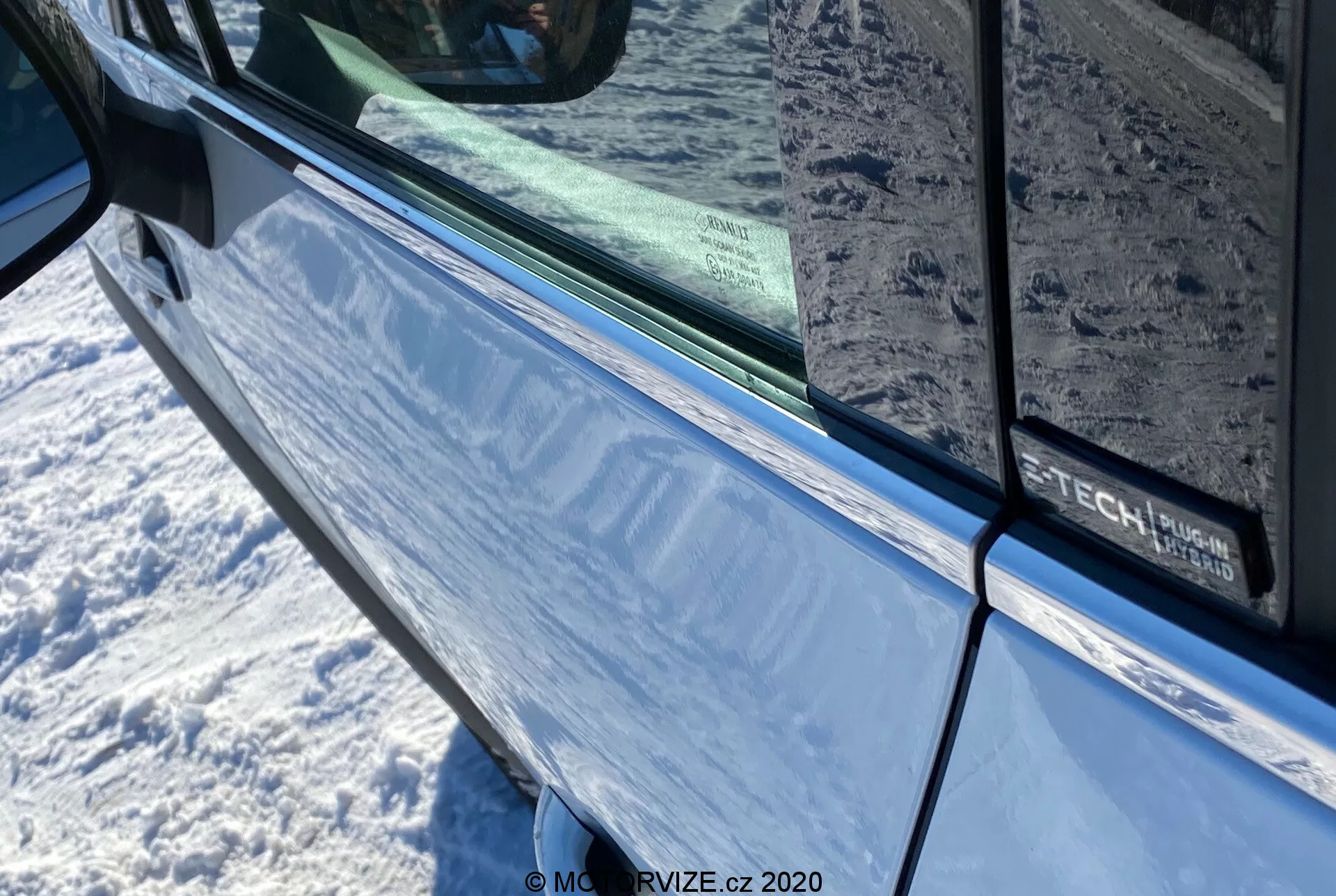 Primo piano della Renault Captur 2020 in blu metallizzato focalizzato sulla porta del conducente e sullo specchietto laterale con freccia integrata. L'immagine mette in evidenza l'area della porta con un bordo nero lungo il bordo della finestra tinta, riflettendo l'ambiente circostante. Sulla porta è parzialmente visibile un marchio che indica un possibile pacchetto di tecnologia o allestimento.