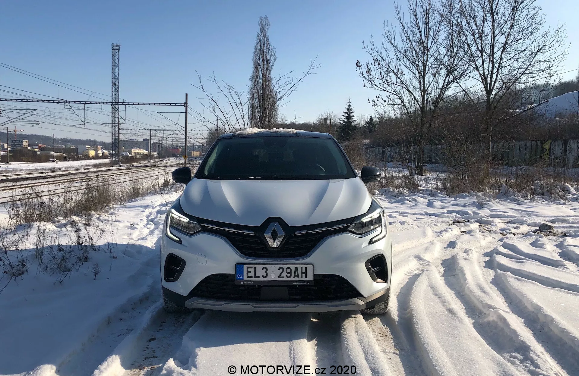 Vista anteriore diretta del Renault Captur 2020 su una strada coperta di neve, che mostra in dettaglio la griglia anteriore, i fari, il cofano scolpito e il paraurti anteriore durante il giorno. Sono visibili le tracce degli pneumatici, segno di movimenti recenti. Il design del veicolo suggerisce uno stile aerodinamico, con un emblema del marchio prominente e un sistema di illuminazione avanzato. Possibili sensori o fotocamere di assistenza alla guida potrebbero essere integrati nel paraurti anteriore.