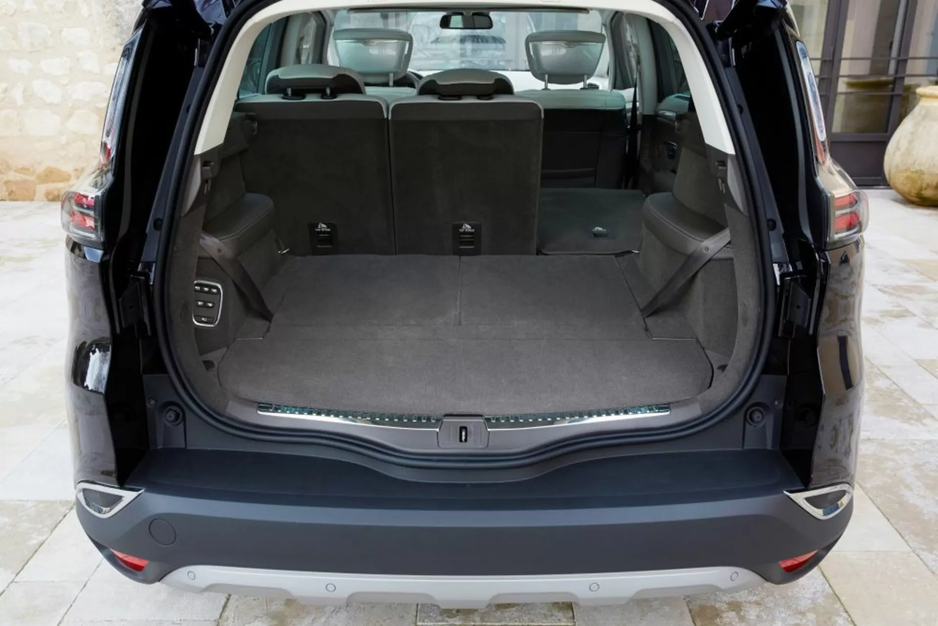 Vista interna posteriore di una Renault Espace 2015 che mostra un'area di carico pulita con il portellone aperto, dotata di un pavimento di carico piatto, sedili posteriori con divisione 60/40, una presa di corrente, ganci per il carico, pulsante di controllo per il portellone elettronico, e una striscia decorativa cromata, sotto illuminazione naturale.